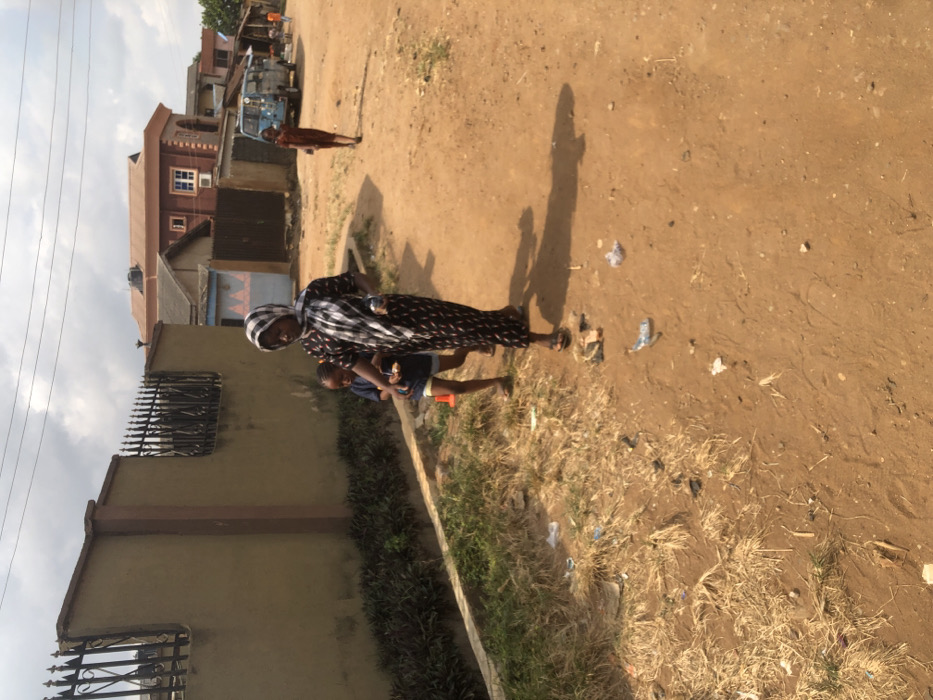 A daytime scene depicts an unpaved, reddish-brown dirt road in Egan, Nigeria, under an overcast sky. Three individuals are visible. In the foreground, an adult female wearing a patterned headscarf and a dark, patterned long dress walks towards the viewer's right, casting a prominent shadow. She wears sandals and holds an object in her left hand. A young child, wearing a dark blue shirt and light shorts, walks slightly behind and to the left of the adult, holding a light-colored object. An orange traffic cone is present on the ground near this child. To the left of the road, a low concrete curb borders a strip of green grass and dry vegetation. Beyond this, a tall, tan concrete wall with dark metal spiked fencing on top runs parallel to the road. Scattered litter, including plastic wrappers, is visible on the dirt road and in the vegetation. In the mid-ground, a second child, dressed in reddish-brown attire, walks further down the road on the right side. A light blue van or small bus is parked or stationary further down the road. Various residential buildings, including a two-story structure with a reddish-brown roof, are visible in the background. Overhead power lines traverse the sky.