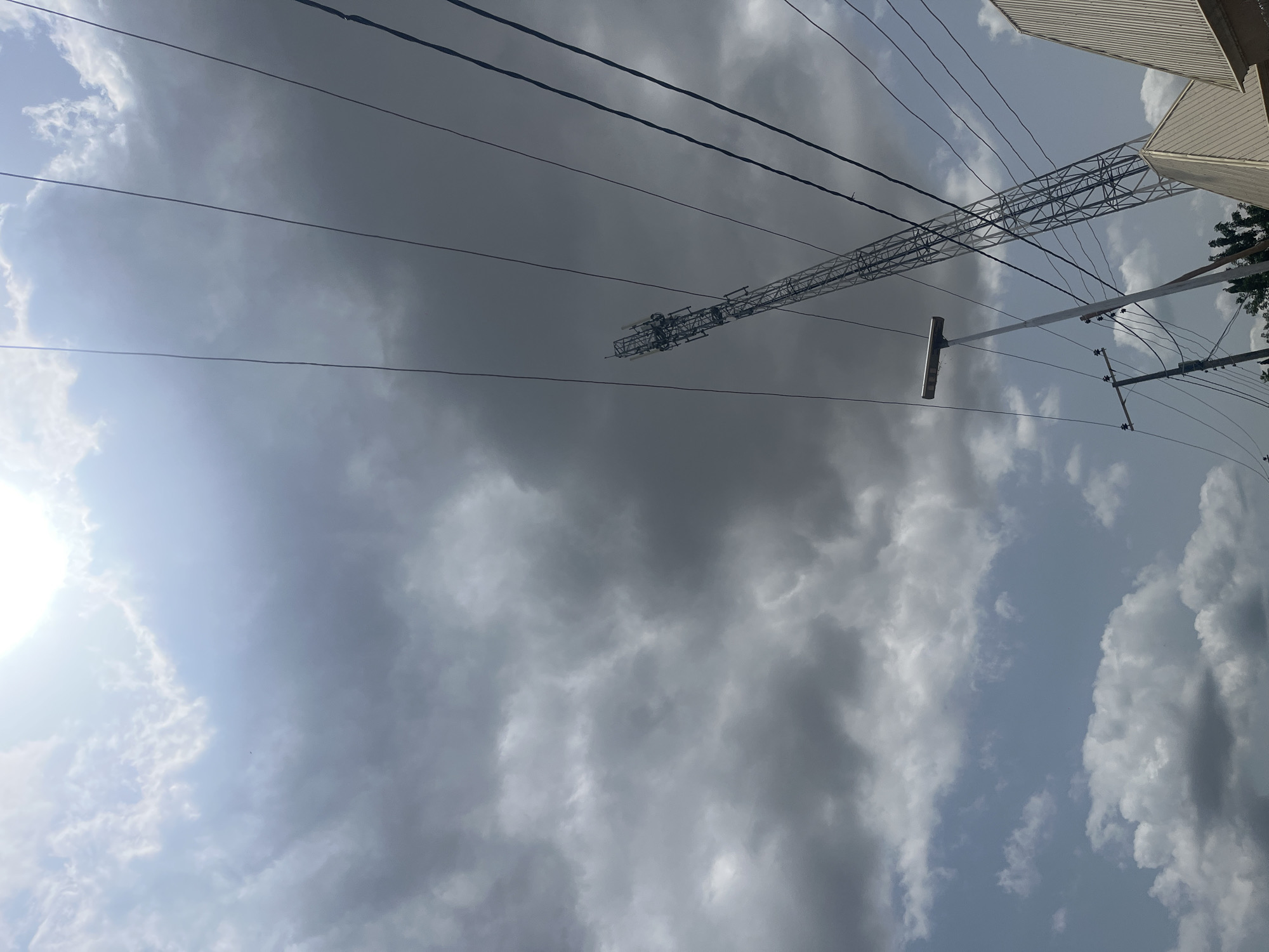 The image depicts a sky with dramatic cloud formations and a bright sun peeking through. In the foreground, a cell tower looms large, crisscrossed by power lines. A street light stands tall to the left of the tower, and a building with a ribbed roof is visible in the lower right corner. The scene appears to be captured from a low angle, looking upwards. The lighting suggests it's daytime, possibly late afternoon due to the angle of the sun and the presence of shadows. The environment is urban or semi-urban, characterized by infrastructure like towers and power lines. There are no people visible in the frame.