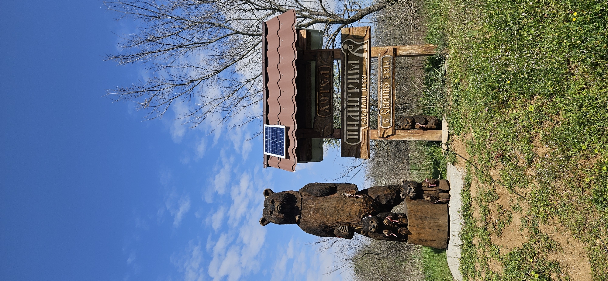 The image depicts a wooden sculpture of a bear with cubs located near a sign in Dobrovo, Bulgaria. The sculpture stands on a grassy area adjacent to a walking path. The sign has a brown metal roof with a solar panel. There are words in Bulgarian written on the sign. The sky is blue with scattered clouds. Bare tree branches are visible in the background.