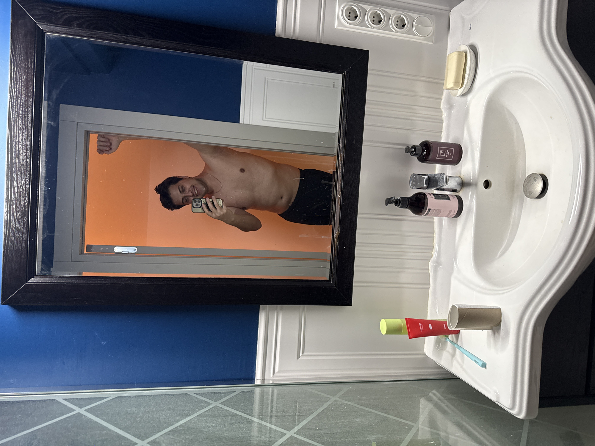 The image shows a bathroom scene, possibly located in an apartment in Vidnoye, Russia, based on the style of fixtures. A black-framed mirror hangs on a blue wall above a tiled floor. Reflected in the mirror is a bare-chested man holding a smartphone, taking a photo of himself in front of an orange wall. To the right of the mirror, a white sink is mounted on the wall. On the sink are three electrical outlets, soap in a dish, two bottles of soap/lotion, a faucet, and a drain. Below the sink, are items including a tube of toothpaste, an empty toilet paper roll, and a toothbrush.