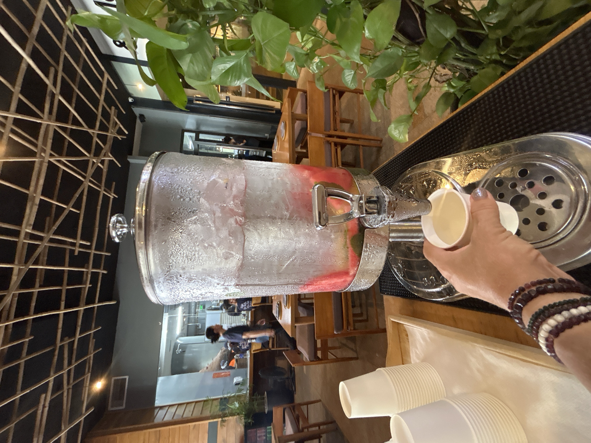 In Nha Trang Ward, Vietnam, a hand wearing multiple bracelets is pouring a beverage from a large dispenser into a white paper cup. The dispenser, containing ice and fruit, is positioned on a counter next to a stack of paper cups. The surrounding environment includes indoor plants and furniture within a commercial establishment.