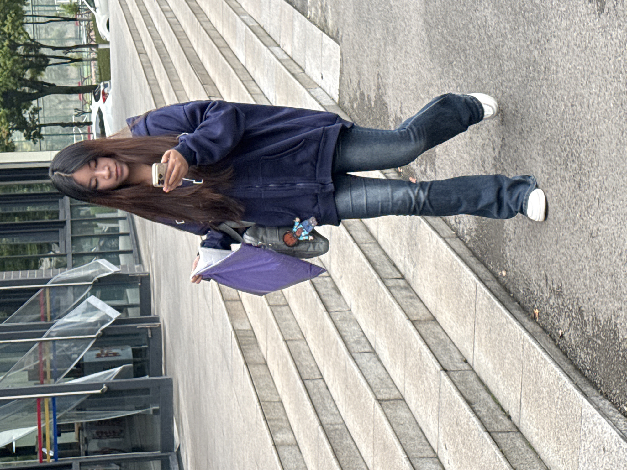 A young woman is seen on a stepped concrete area in what could be Jiangjin, China. She is holding a smartphone, possibly interacting with it. The woman is wearing jeans, a dark blue zip-up hoodie, and white sneakers. She has long dark hair. A grey bag and a purple object, possibly an umbrella, are in her possession. A modern building with glass windows and decorative elements is visible behind her. A few vehicles are further in the background, and some trees are also visible. The weather appears to be overcast.