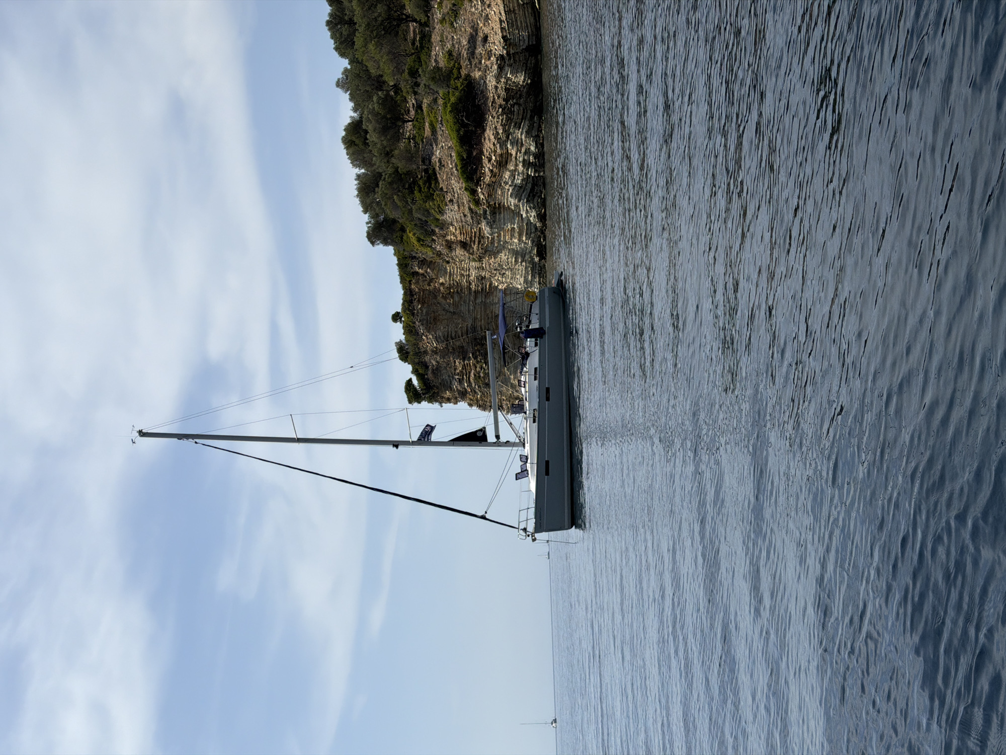 A sailboat is anchored in calm, blue water near a rocky, tree-covered cliff face. The sky is partly cloudy with a light blue hue. The sailboat is gray with a tall mast and appears to be unoccupied. In the distance, to the left, two other smaller sailboats are visible. The scene is serene and captures a typical Mediterranean coastal landscape. This image likely depicts a location within the Municipal Unit of Kastos, Greece, known for its clear waters and scenic islands.