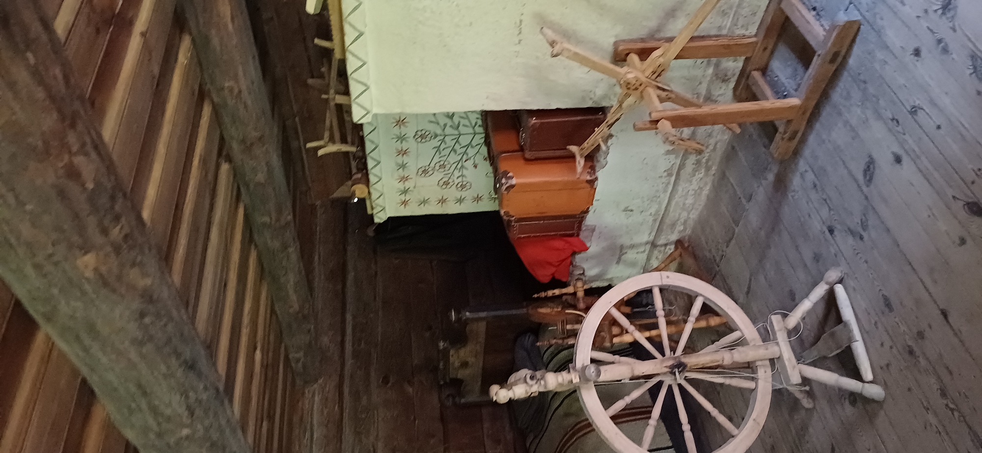The image shows the interior of a rustic room, possibly a cabin or an old house. The ceiling is made of wooden planks supported by thick, rough-hewn logs. The walls are also wooden, with visible grain and texture.
In the foreground, to the left, is a wooden spinning wheel with a large spoked wheel and a spindle mechanism. The wood appears light-colored and worn. Behind the spinning wheel, a bed with striped bedding is partially visible.
To the right, there is a white stove or hearth, decorated with painted floral patterns and star-like motifs in green, red, and white. Stacked on top of or next to the stove are several vintage suitcases in shades of brown and orange. A red cloth is draped over the suitcases.
Further to the right, another wooden contraption, possibly another type of spinning or weaving tool, is visible. It is made of lighter wood and has a complex arrangement of arms and wheels.
The floor is made of wooden planks, showing signs of age and wear, with dark knots and grain patterns. The overall impression is one of a historical or rural setting, filled with antique domestic items. There are no people visible in the image. The lighting is somewhat dim, suggesting an indoor space without direct sunlight.