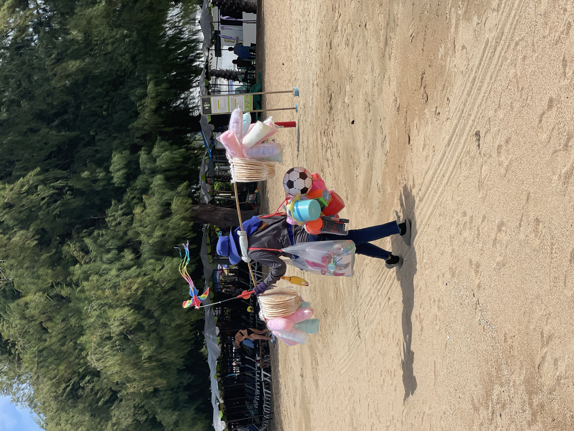A person wearing a blue hat, a gray long-sleeved shirt, and blue pants walks along a sandy beach in Pattaya City, Thailand. They are carrying a pole over their right shoulder, from which various items are suspended. On the left side of the pole, several pastel-colored cotton candy puffs and what appear to be paper lanterns are hanging. On the right side, a black and white soccer ball, colorful plastic buckets, and a yellow toy are attached. A kite with rainbow-colored streamers is held up by a stick, also attached to the pole. In the background, numerous beach umbrellas and lounge chairs are set up. A woman in a black bikini is visible near the umbrellas. The ground is sandy with some tire tracks visible. The sky is bright and the sun is casting a shadow of the person onto the sand.