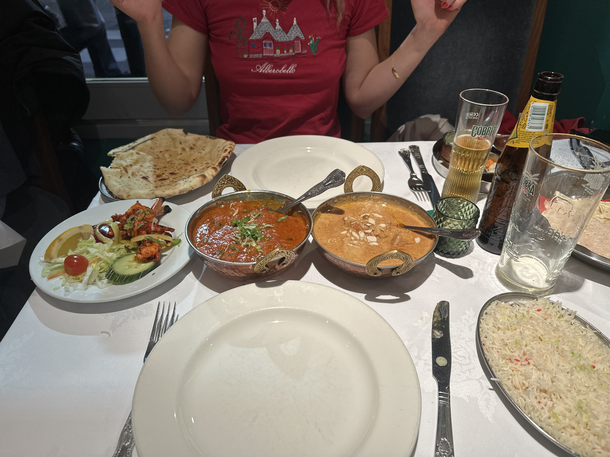 The image shows a table laden with Indian food at a restaurant. Two dishes of curry, one orange-red and one tan with almond slivers, sit in ornate metal bowls. There is naan bread, a plate of tandoori shrimp with a small salad, and a dish of rice. Two empty white plates are set, one for each person, and silverware is present. Two Cobra beer bottles and two matching glasses are visible, one glass filled with beer. A woman in a red "Alberobello" t-shirt is seated at the table, her hands raised in a gesture. The location appears to be a restaurant, likely in London, UK.