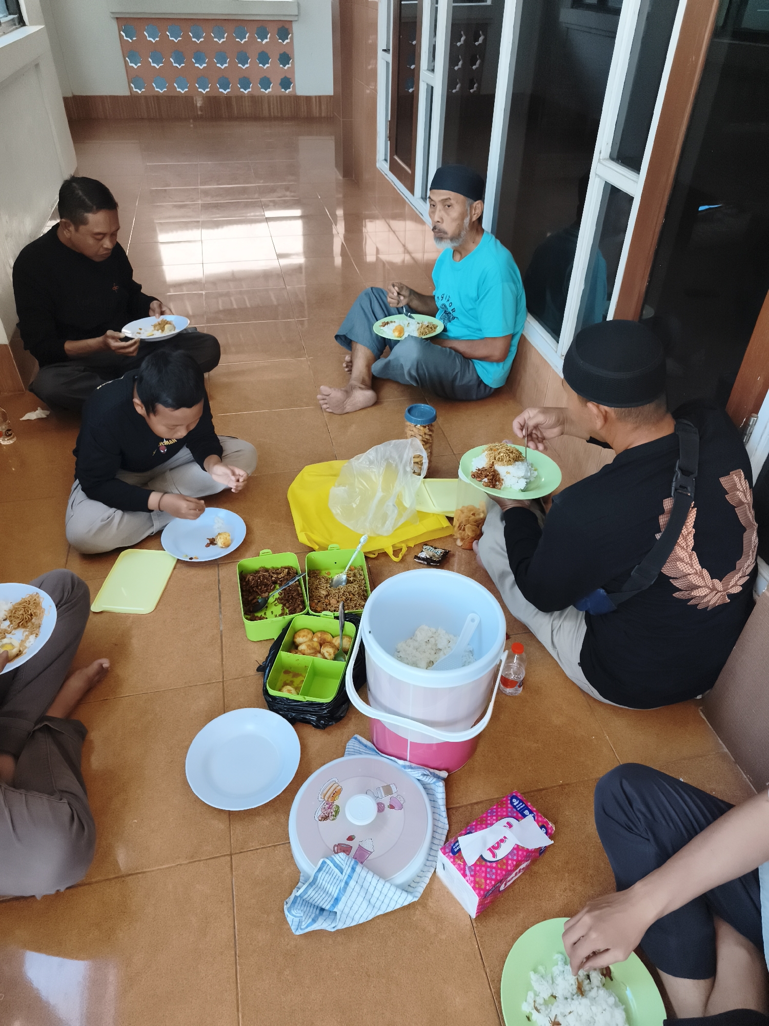 The image depicts a group of people sitting on a tiled floor, likely outdoors or in a covered patio area, in Tonasa, Indonesia, enjoying a meal. There are at least five individuals visible, dressed in casual attire. The scene is informal, with food spread out on the floor.
In the foreground, to the left, a person's legs are visible, with a plate of noodles and eggs in front of them. Next to this, a person is seated, wearing dark pants and a black long-sleeved shirt, with a plate of food. Further to the left, another person in a black long-sleeved shirt and grey pants is also seated, holding a plate.
In the mid-ground, a young person in a black long-sleeved shirt and grey pants is sitting cross-legged and appears to be eating. In front of this person is a blue plate, and a light green tray. To the right of the young person, two green food containers are visible, one filled with a noodle-like dish and the other with small, round, cooked items, possibly eggs. A large white bucket with a pink lid, containing rice, is also present, along with a small bottle of sauce.
In the background, an older man wearing a light blue t-shirt and dark pants sits with a plate of food. Another individual in a black cap and dark clothing is seated further back, also with a plate.
The environment appears to be a tiled patio with a patterned wall in the background and large windows or glass doors. The lighting suggests it is daytime, with sunlight casting reflections on the floor. A yellow plastic bag is also visible near the food containers. The overall impression is a communal and relaxed meal.