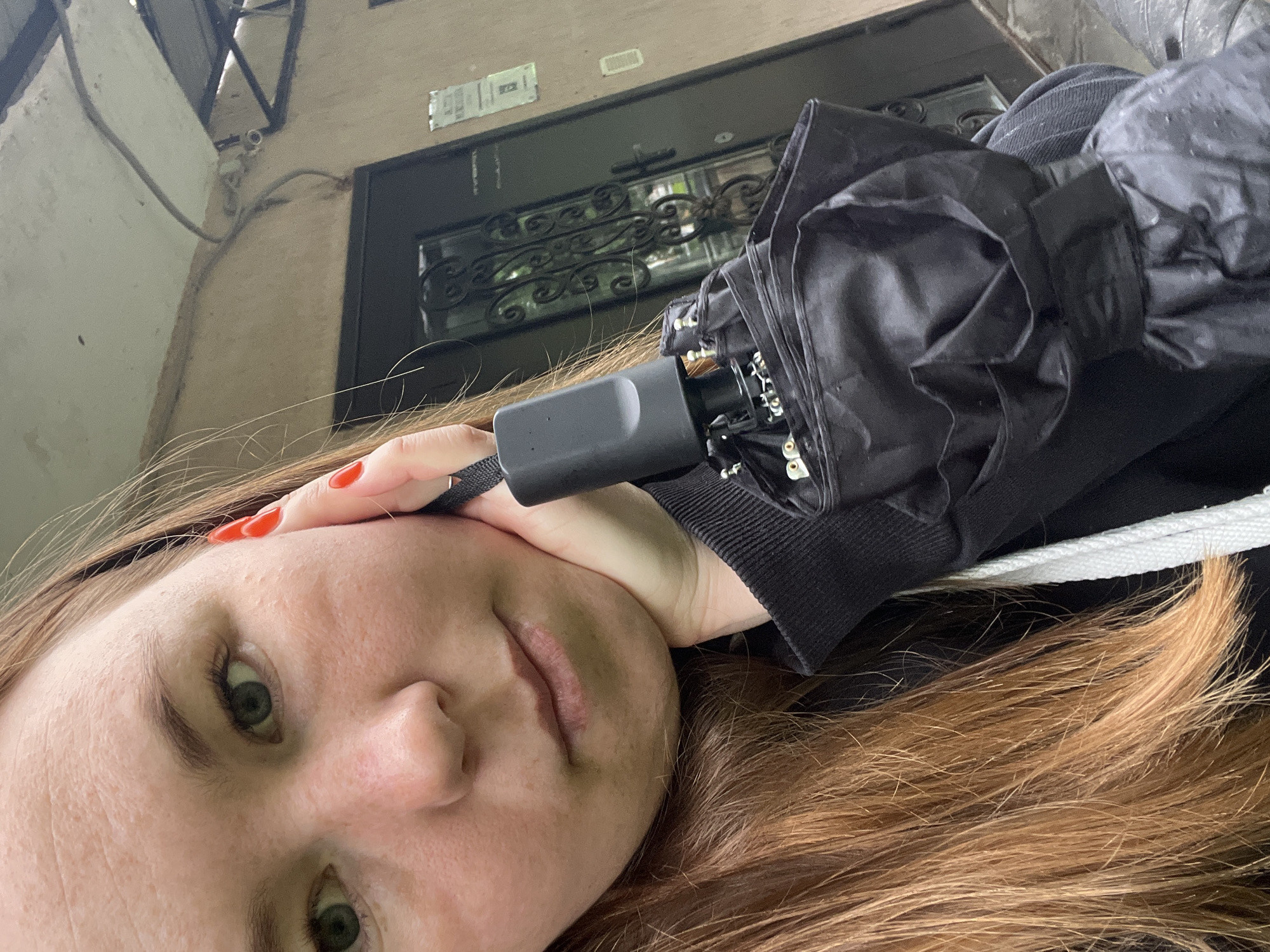 The image, taken with a tilted perspective, features a young woman in a selfie. She has light brown to auburn hair, fair skin, and green or hazel eyes, with a neutral expression. She wears red nail polish on her visible thumb and holds a black folded umbrella by its handle, resting it against her head. She is dressed in a black jacket or hoodie, with a white drawstring visible near her shoulder.
The background depicts the exterior of an old building. A dark door with ornate black metal grillwork in its window section is prominent. Above the door, a light-colored rectangular sticker or sign displays some illegible text. Wires are visible running along the building's light-colored wall. A small white device, possibly a security camera or sensor, is attached to the wall above the door. The setting is outdoors during the daytime, and the presence of the umbrella suggests recent or impending rain. The location context is Chaikovsky, Russia.