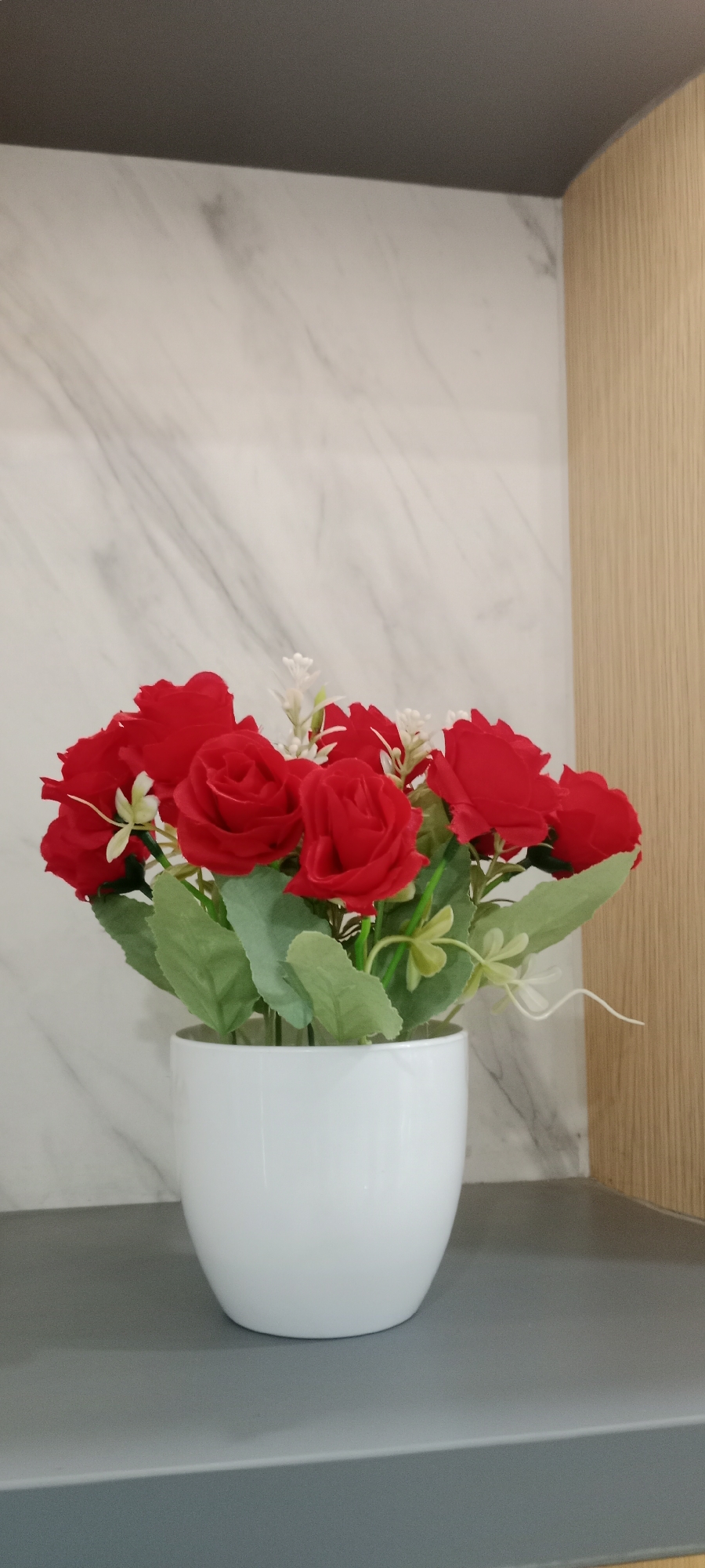 A white ceramic pot holds a bouquet of artificial red roses with green leaves and small white accents. The pot sits on a gray surface, in front of a backdrop of a marble-patterned wall on the left and a light brown wooden panel on the right. The image appears to be an indoor scene, possibly a decorative element in a home or establishment in Tuban, Indonesia.
