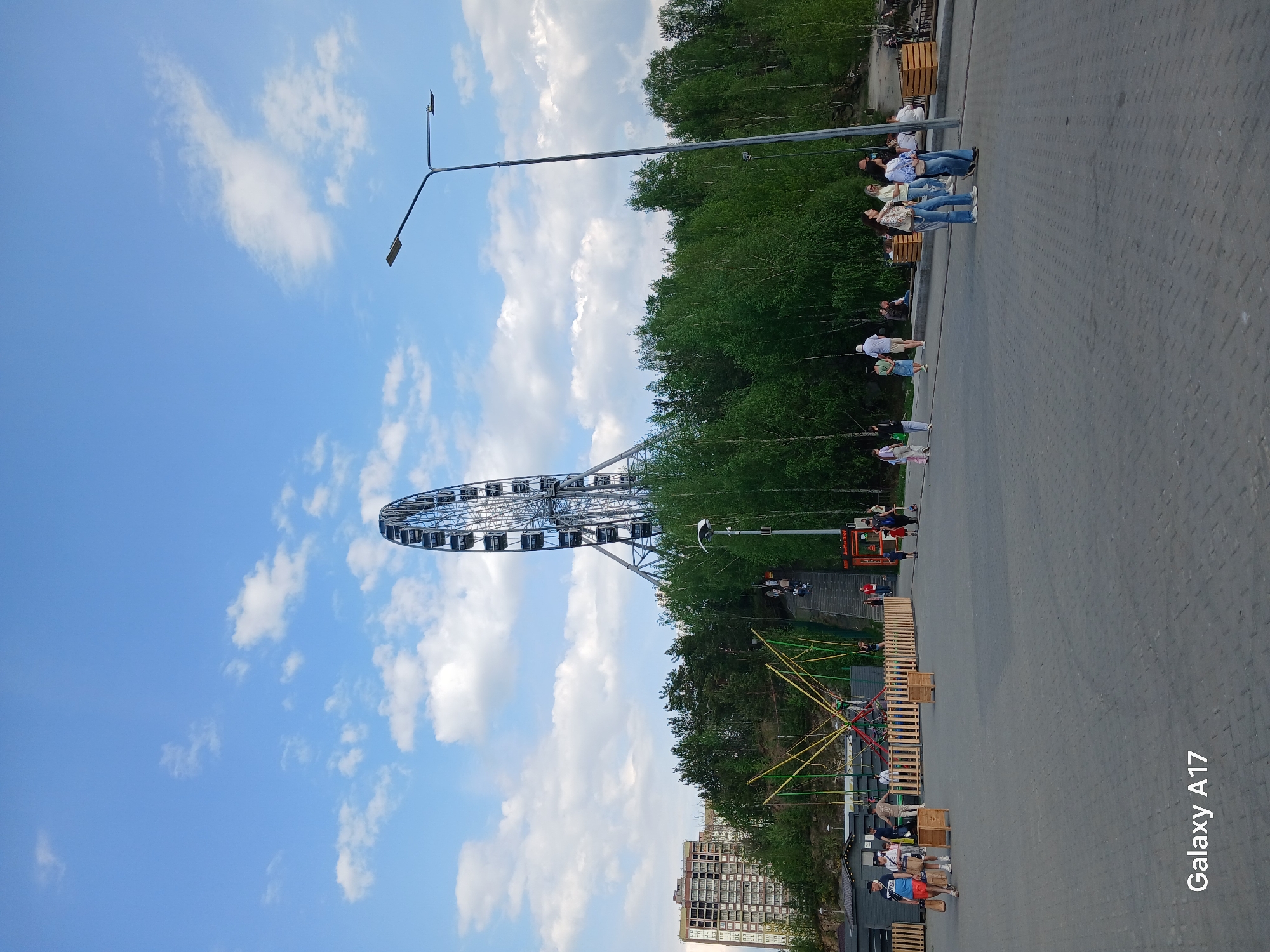 The image captures a Ferris wheel on a cloudy day in what appears to be a park in Yekaterinburg, Russia. The wheel is a large silver structure with passenger cars. In the foreground, there is a paved area with people walking and sitting on benches. A street light is visible. A wooden trampoline-like structure is near the bottom of the frame. There is a building in the very bottom of the frame, and green trees are in the background.