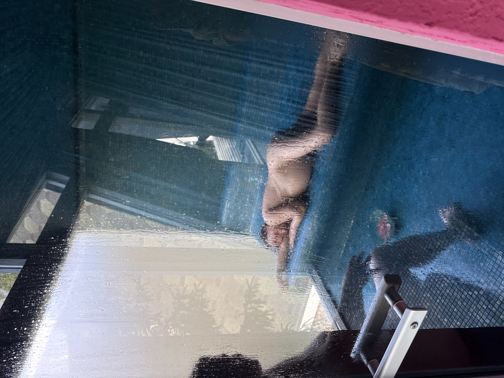 The image shows a blurred reflection of a person in a window. The person appears to be lying on a blue surface. Outside the window, there are glimpses of buildings and greenery. The window has a handle. The upper frame of the window is painted pink. The image location is Чёрное, Russia.