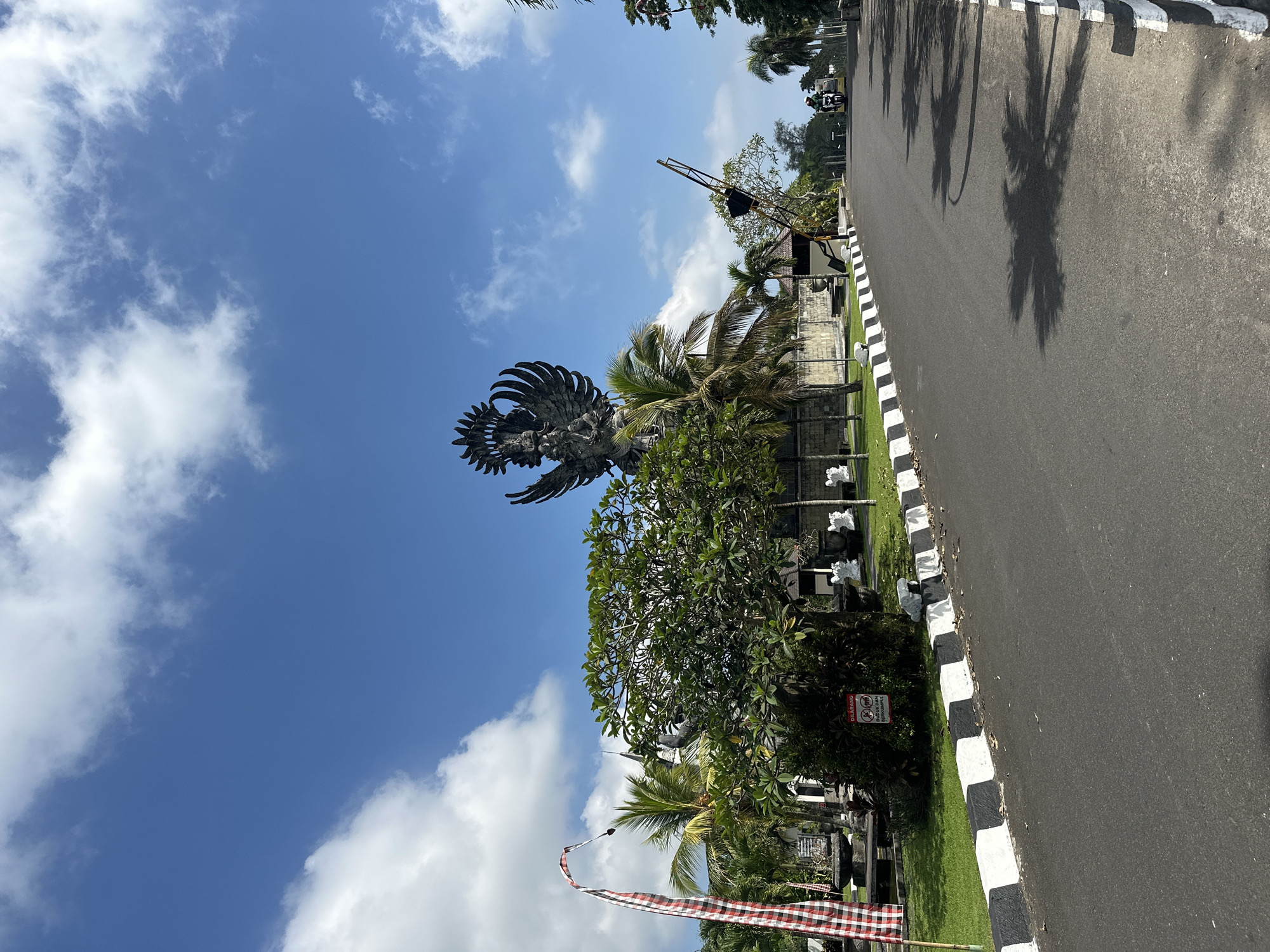 The image depicts a large statue of a bird-like figure, possibly Garuda Wisnu Kencana, in Kuta Selatan, Bali, Indonesia. The statue stands against a blue sky with scattered clouds. Palm trees and other greenery surround the statue. A road runs along the right side of the image, with a black and white striped curb. Shadows of palm trees fall across the road. A red and white checkered flag is visible near the base of the statue.