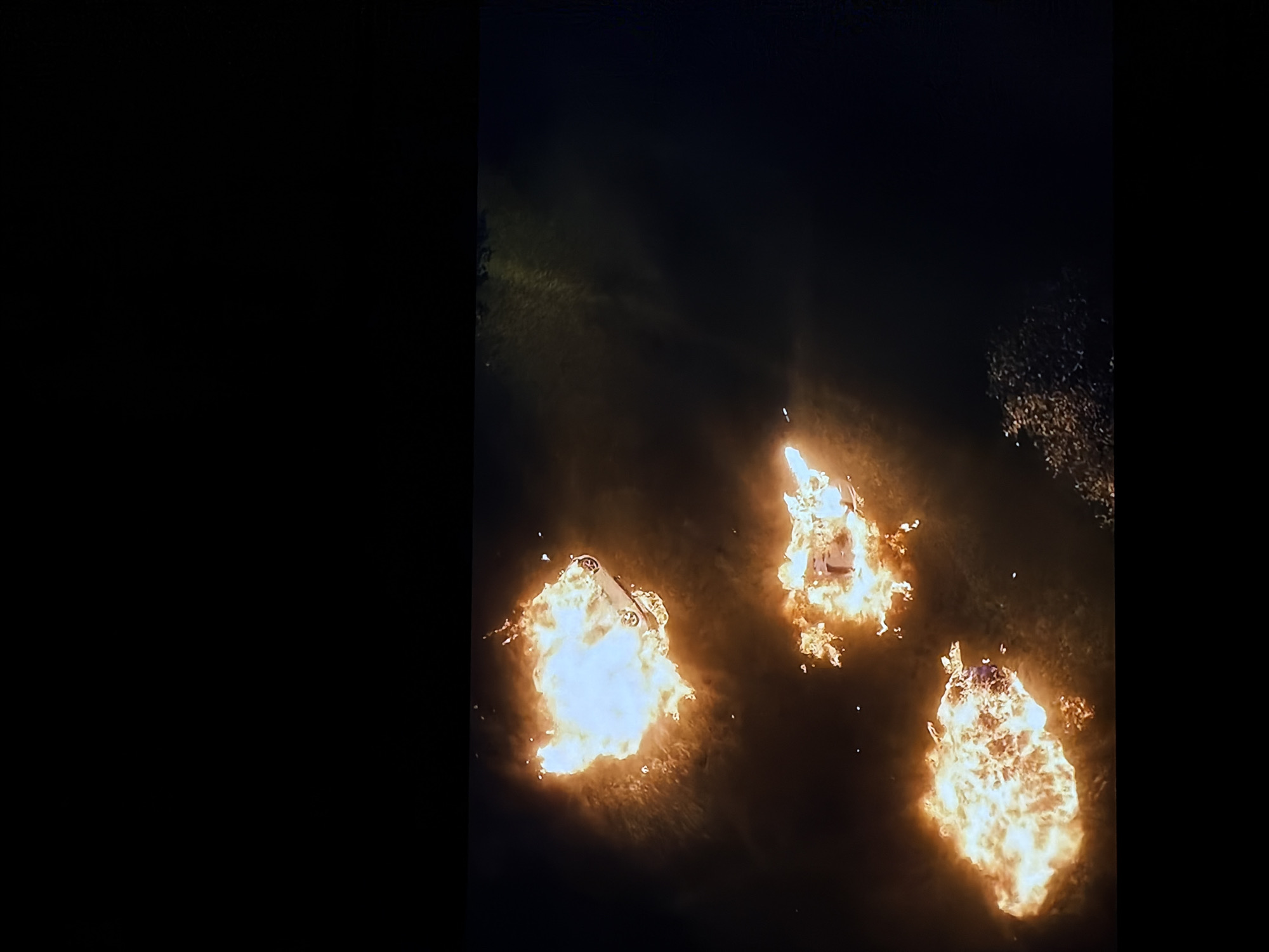 The image displays a nighttime scene in Sofiivska Borshchahivka, Ukraine, dominated by three large, bright fires. Each fire appears to engulf a vehicle, with the distinct shapes of cars discernible within the intense flames. The fires cast a strong orange and yellow glow against the otherwise dark background, illuminating patches of what appears to be an outdoor, possibly unpaved or grassy area. No people are visible in the frame. The scene suggests a significant destructive event involving burning vehicles. The time of day is night, emphasized by the darkness and the fires acting as the primary light sources. No text is visible in the image. The overall impression is one of destruction and a serious incident.