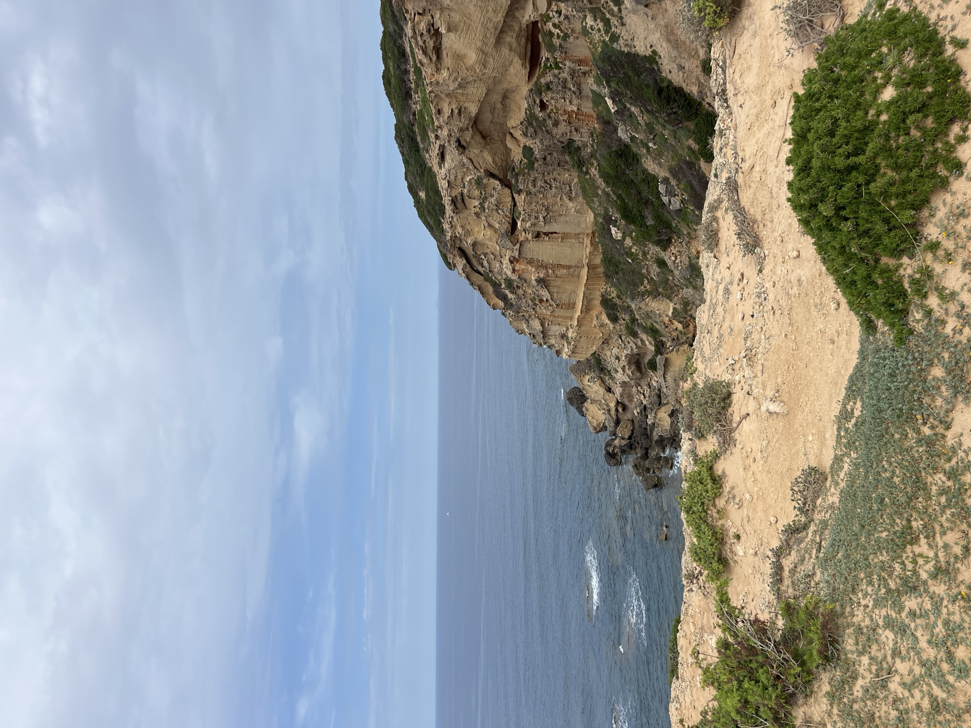 The image shows a rocky coastline near San Vero Milis, Italy. A steep cliff dominates the right side of the frame, featuring layers of tan and brown rock formations with patches of green vegetation. The cliff drops into a dark blue sea that occupies the lower center of the image. The left side of the image is filled with a light blue sky with thin, wispy clouds. The scene is calm, with no visible human activity or structures.