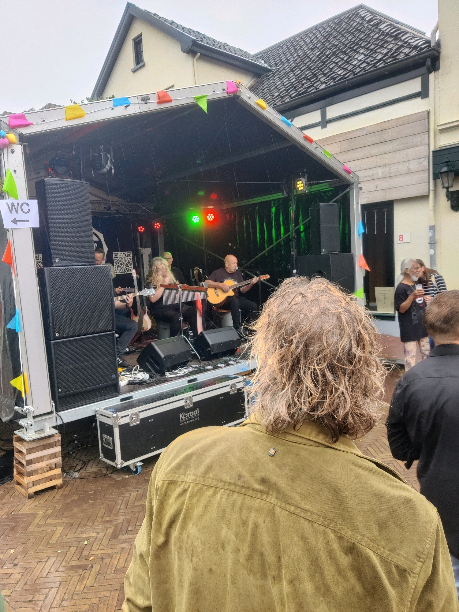 The image shows an outdoor musical performance on a stage set up on a trailer. The stage has a black backdrop with green drapes and is illuminated by colored stage lights. Three musicians are visible on the stage, two playing guitars and one playing a stringed percussion instrument. A small crowd of people is gathered to watch the performance, with the back of a person with long, wavy blonde hair prominently in the foreground. The event appears to be taking place in a street in Winterswijk, Netherlands, suggested by the architectural style of the buildings in the background and the presence of a sign with "WC" and an arrow, commonly seen at public events in European cities. The weather appears to be overcast, with some indication of rain on the stage. The overall scene depicts a community event or festival.