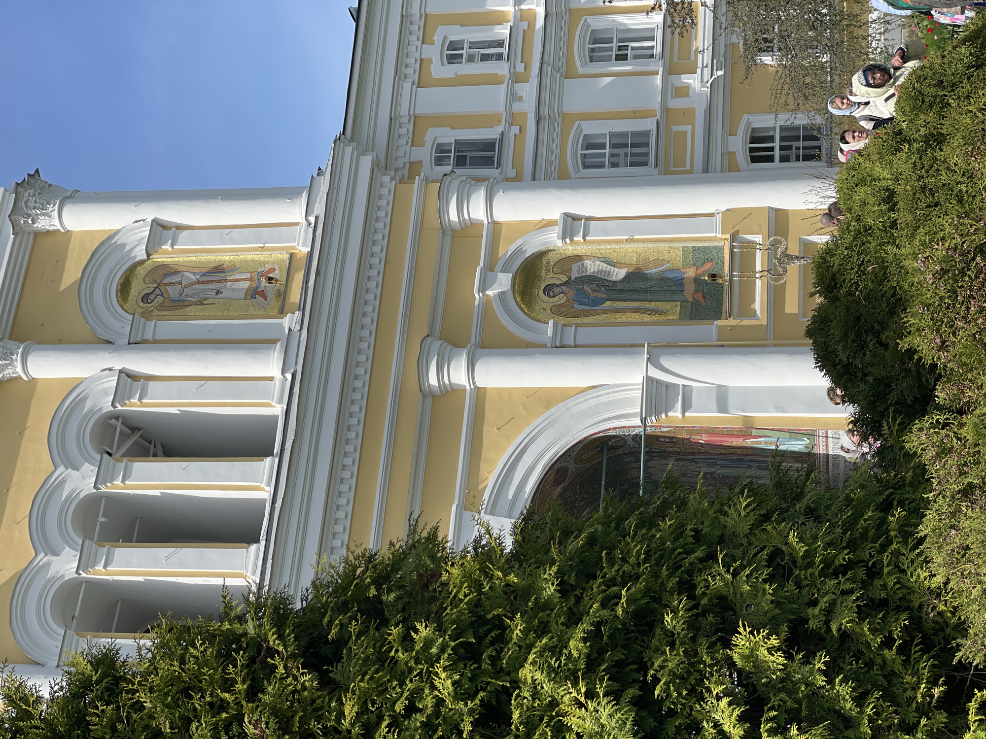 The image depicts the exterior of a building in an unknown location, city Unknown, Unknown, with yellow and white architectural details. There are two visible mosaic icons adorning the facade. A large evergreen bush partially obscures the lower portion of the building. A small group of people is seen in the background, partially obscured by the bush. The sky is clear and blue.
