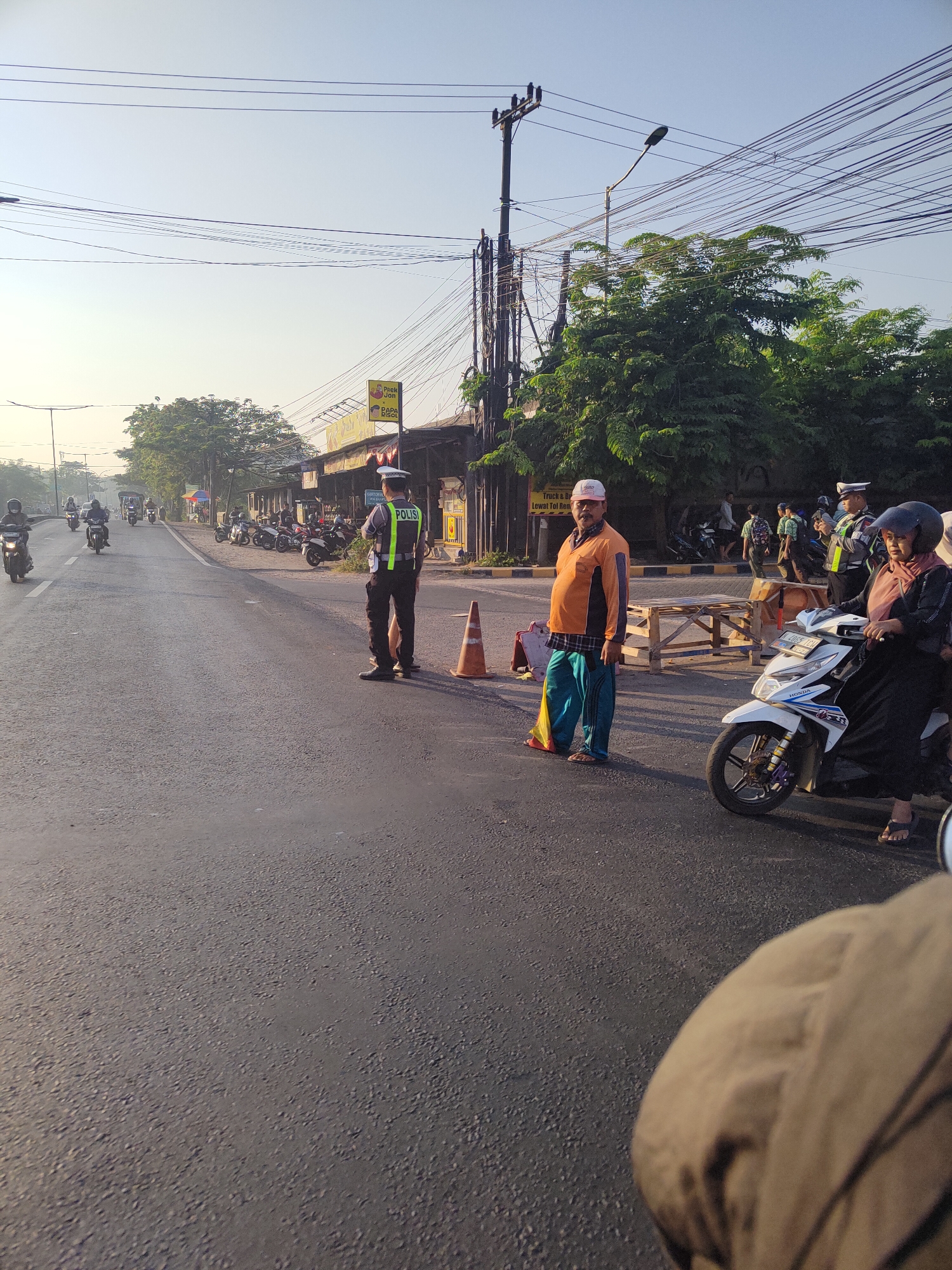 The image depicts a street scene in Raci, Indonesia, during daylight hours. A traffic police officer in uniform, identified by the "POLISI" vest, stands on the right side of the road, facing away from the camera. The officer is wearing a dark uniform with a reflective vest. In front of the officer, an orange traffic cone is placed on the asphalt.
To the right of the police officer, a man in an orange long-sleeved shirt and blue pants stands. He is wearing a white cap and appears to be observing the traffic. Next to him, a woman on a white motorcycle, wearing a black jacket and a hijab, is seated on the vehicle.
The road appears to be a two-lane street with traffic moving in both directions. Several motorcycles are visible on the road and parked on the side. Buildings line the right side of the street, with some storefronts and awnings. Trees and utility poles with numerous wires are also present in the background. The sky is clear and bright, suggesting a sunny day.
The perspective of the image is from a low angle, possibly from a vehicle on the road, as the foreground shows a blurry brown surface that resembles clothing or a bag. The overall impression is a typical street scene with traffic control and daily activities taking place.