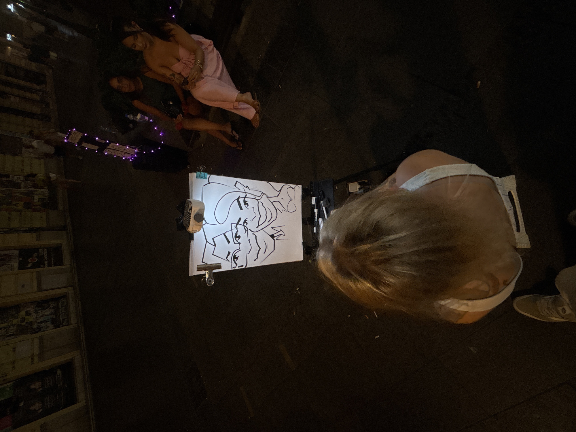 The image depicts a street scene at night, likely in Belgrade, Serbia, with a caricature artist working on a drawing board positioned on a small easel. Two women are seated in front of the artist's setup. The woman on the left wears a dark green top and the woman on the right wears a light pink outfit. The drawing on the board is a caricature of two faces, both wearing masks. In the foreground, a person's blonde hair and shoulder are visible, suggesting they are either the artist or another patron. The background shows a dimly lit street with posters on a building and other people walking by. Decorative purple lights are visible on a stand behind the seated women. The overall atmosphere is one of a lively evening street scene.