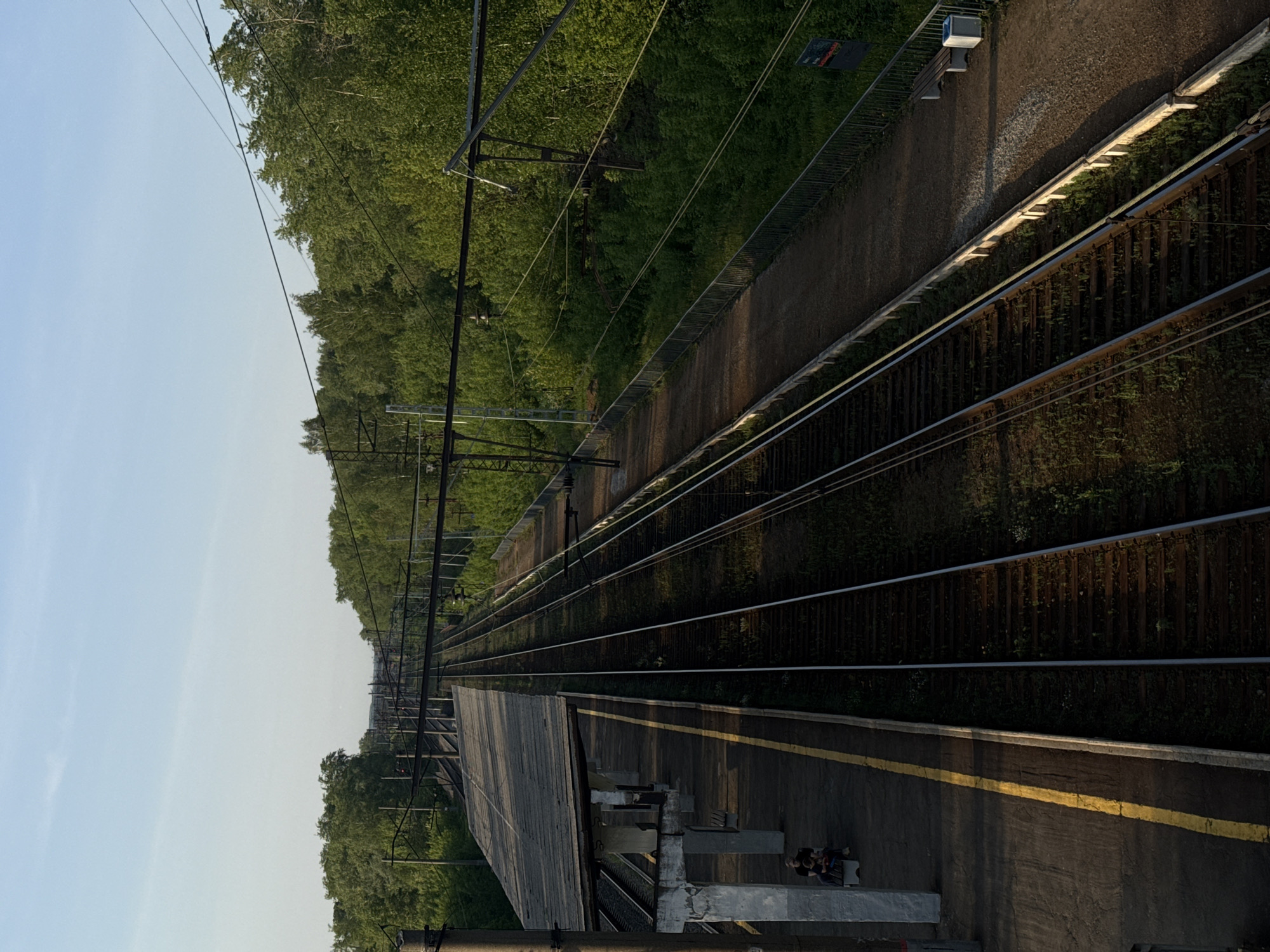 The image shows a view of railway tracks and a platform, likely taken from an elevated position. The tracks extend into the distance, lined by greenery on either side. There are overhead power lines running above the tracks. A platform is visible in the foreground with a yellow safety line painted on the edge. There are two figures, possibly people, standing at the edge of the platform. The image may have been taken in Perm, Russia.