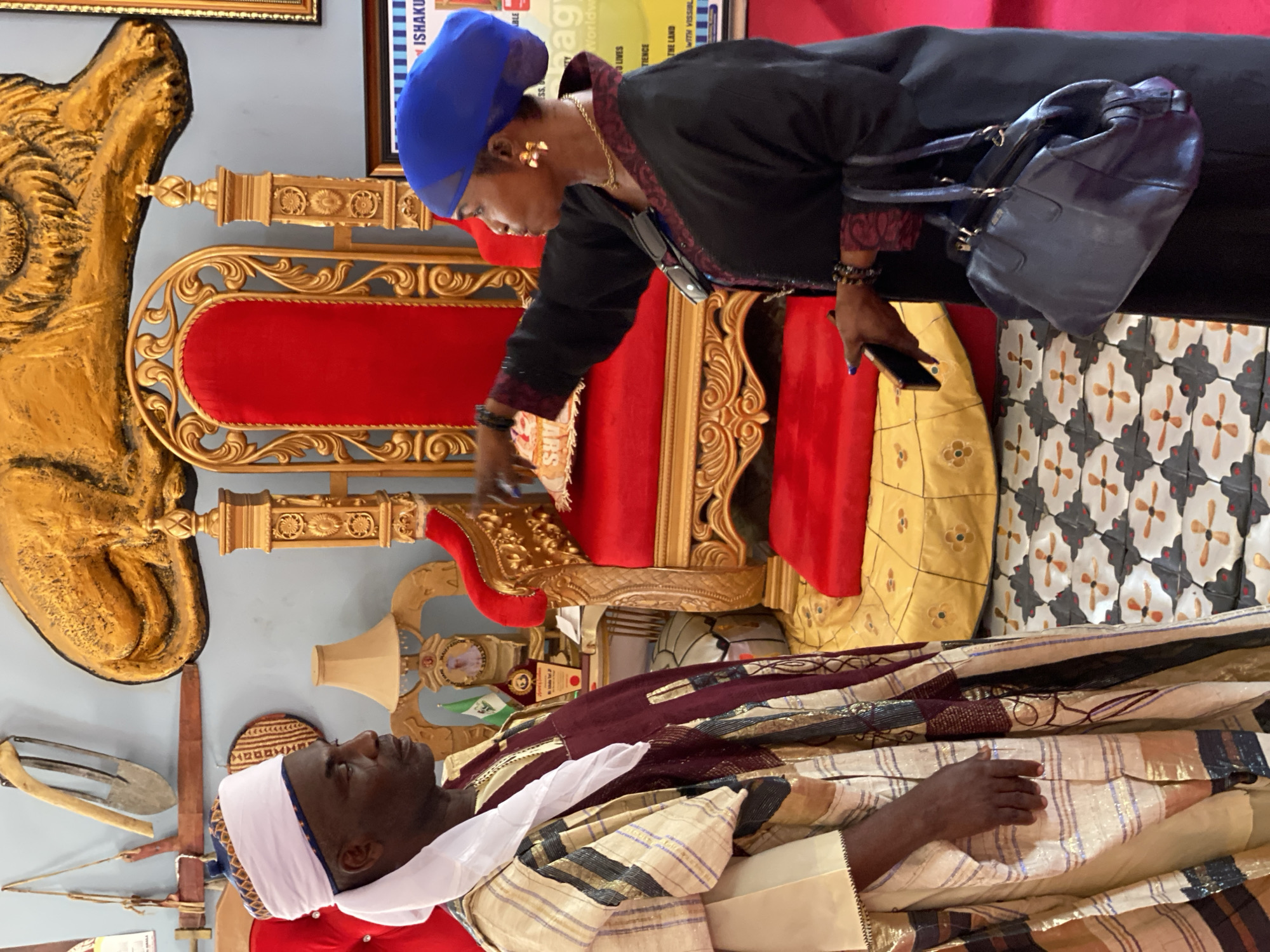 A woman in a black top and blue headscarf, holding a phone and a small container, is interacting with a man lying on a golden throne in an indoor setting, possibly in Sabon Tasha, Nigeria. The man is dressed in a white head wrap and patterned robes. The throne is elaborate, with red upholstery and golden ornamentation. Various objects are on the surrounding walls. The floor has a patterned tile design.