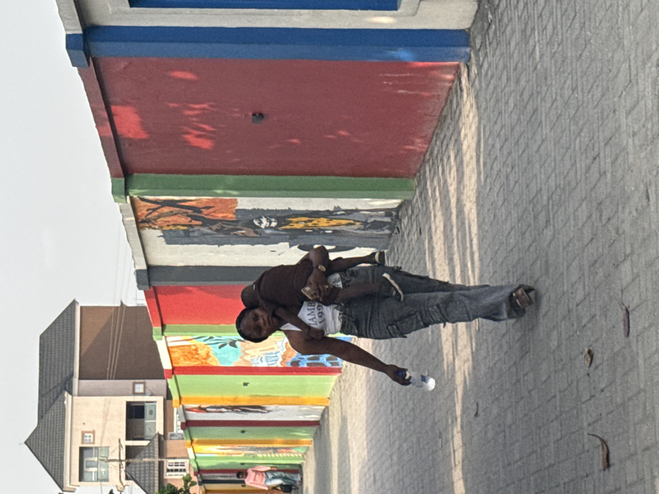 A woman carries a child piggyback style on a pedestrian walkway under a colorful, painted overhang. She wears a white tank top with lettering, gray pants, and sandals. The child wears a brown shirt and dark pants with sneakers. A building is visible on the left side of the frame. The walkway has a brick-like pattern. Sunlight casts shadows on the ground.