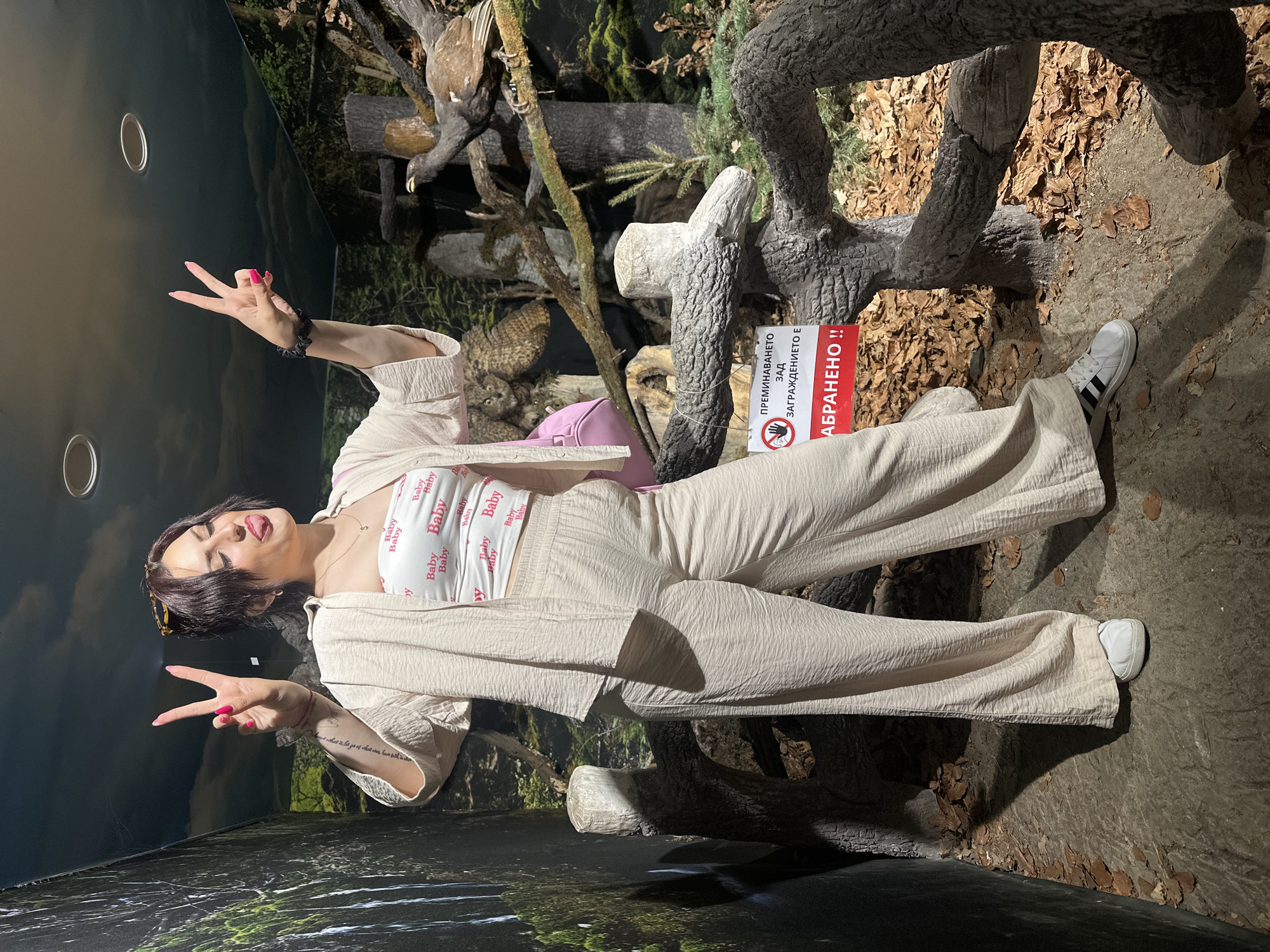 A young woman is posing in a museum display. The museum display appears to be a diorama representing a forest environment, with taxidermied birds and artificial trees. A sign in Bulgarian states "ABRANENO!!", meaning "Forbidden!!". The woman is wearing a white top printed with the word "Baby", and beige pants and jacket. She is making hand gestures. The location is likely the Plovdiv Natural Science Museum in Bulgaria, considering the type of exhibit and the sign in Bulgarian.