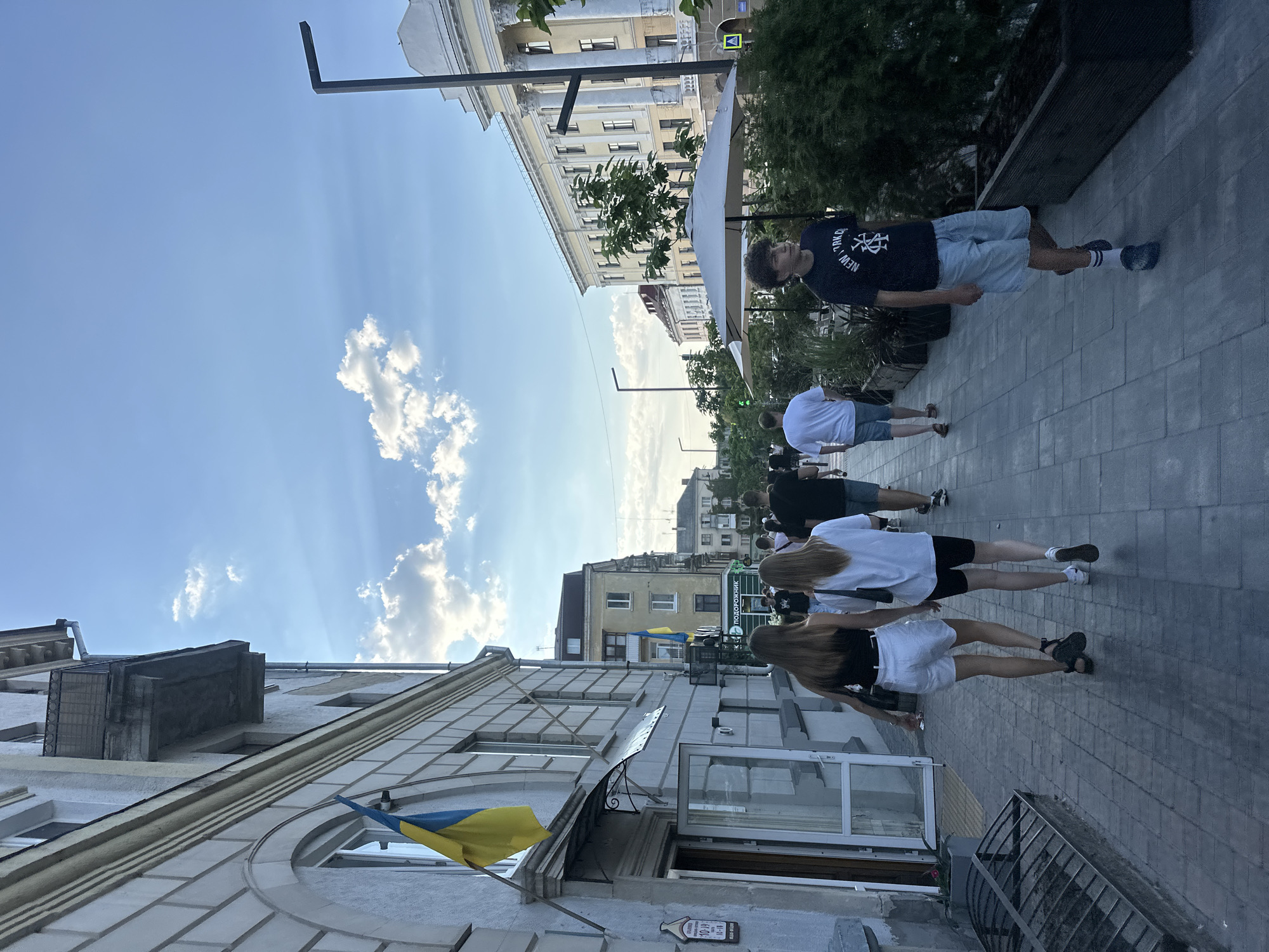 The image depicts a street scene in Ternopil, Ukraine, with people walking along a sidewalk next to a building. A Ukrainian flag is visible on the building's facade. The sky is blue with scattered clouds, and the sun is casting rays through the clouds. Several individuals, including men, women, and possibly teenagers, are walking in the same direction along the sidewalk. Some are dressed in casual summer clothing. A street lamp and trees line the sidewalk. The architecture of the buildings suggests a European city. The overall atmosphere appears to be a pleasant day in a public urban space.