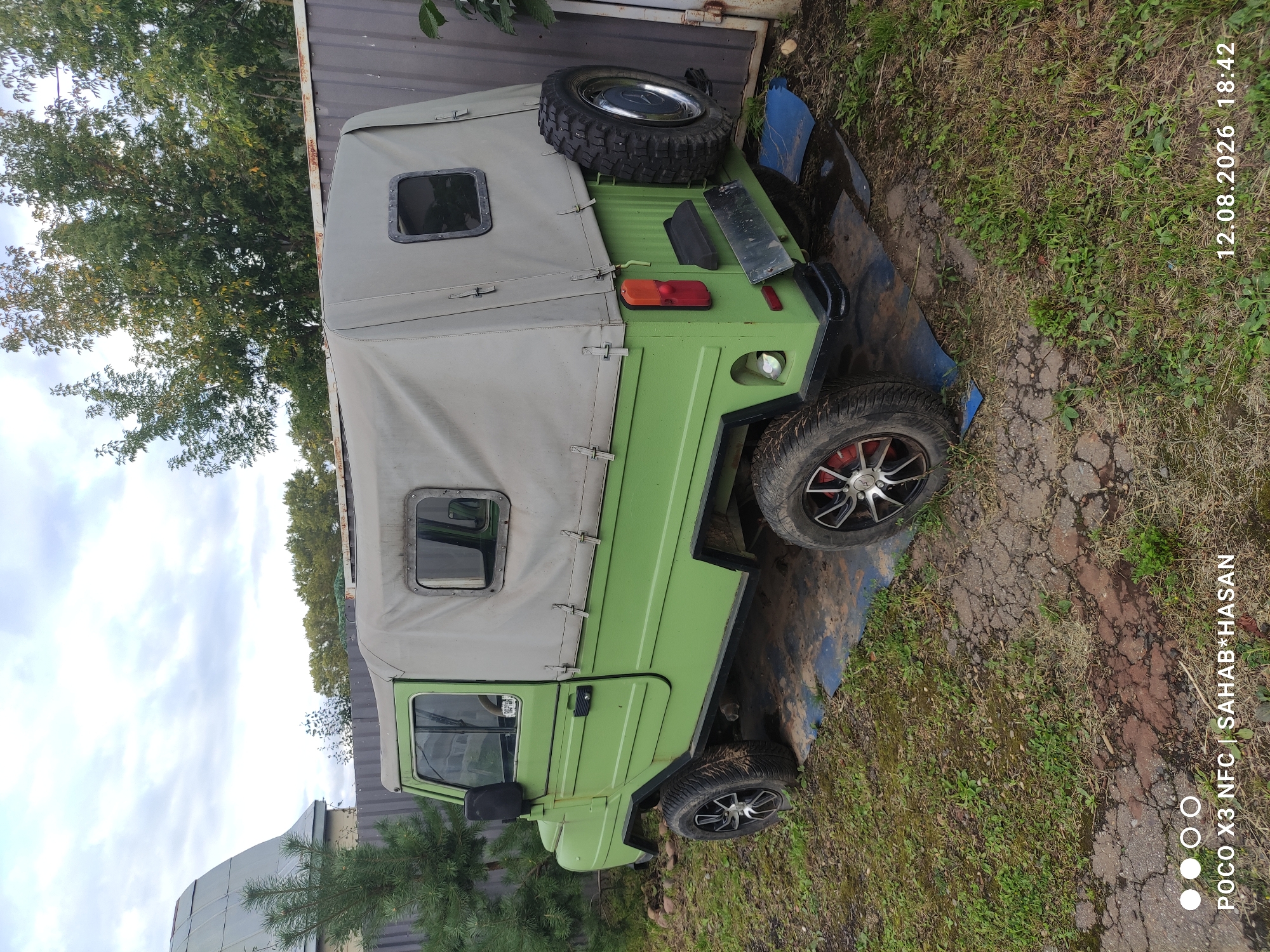 The image shows a green, military-style vehicle parked on cracked pavement and grass. The vehicle has a canvas top, a spare tire mounted on the back, and aftermarket wheels. It is outdoors, with a fence and trees in the background. The sky is overcast. The timestamp at the bottom of the image reads "12.08.2026 18:42," and the text "POCO X3 NFC | SAHAB*HASAN" is also present, indicating the device used for capture and possibly the photographer's name. There is no indication of the location being the city of Kostroma, Russia. There are no people or actions depicted in the image.