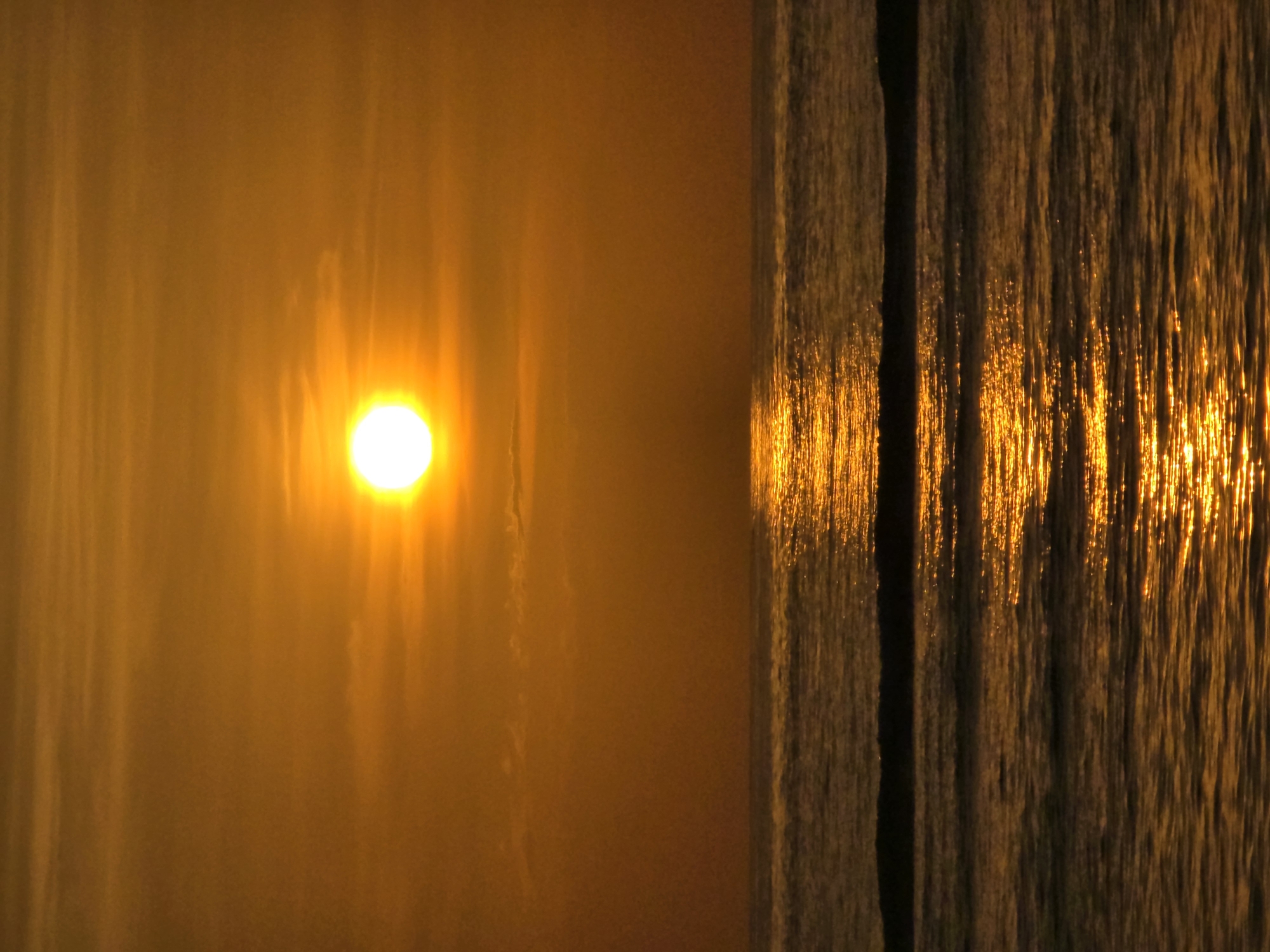 The image shows a sun with rays shining through what appears to be fog. To the right, there is a vertical structure with a gap in the middle, reflecting the sun's light. The vertical structure has a dark, almost wooden-like appearance, and the reflection has a pattern of bright vertical streaks. The location appears to be on a waterfront with the sun shining through fog or mist. The city is unknown.
