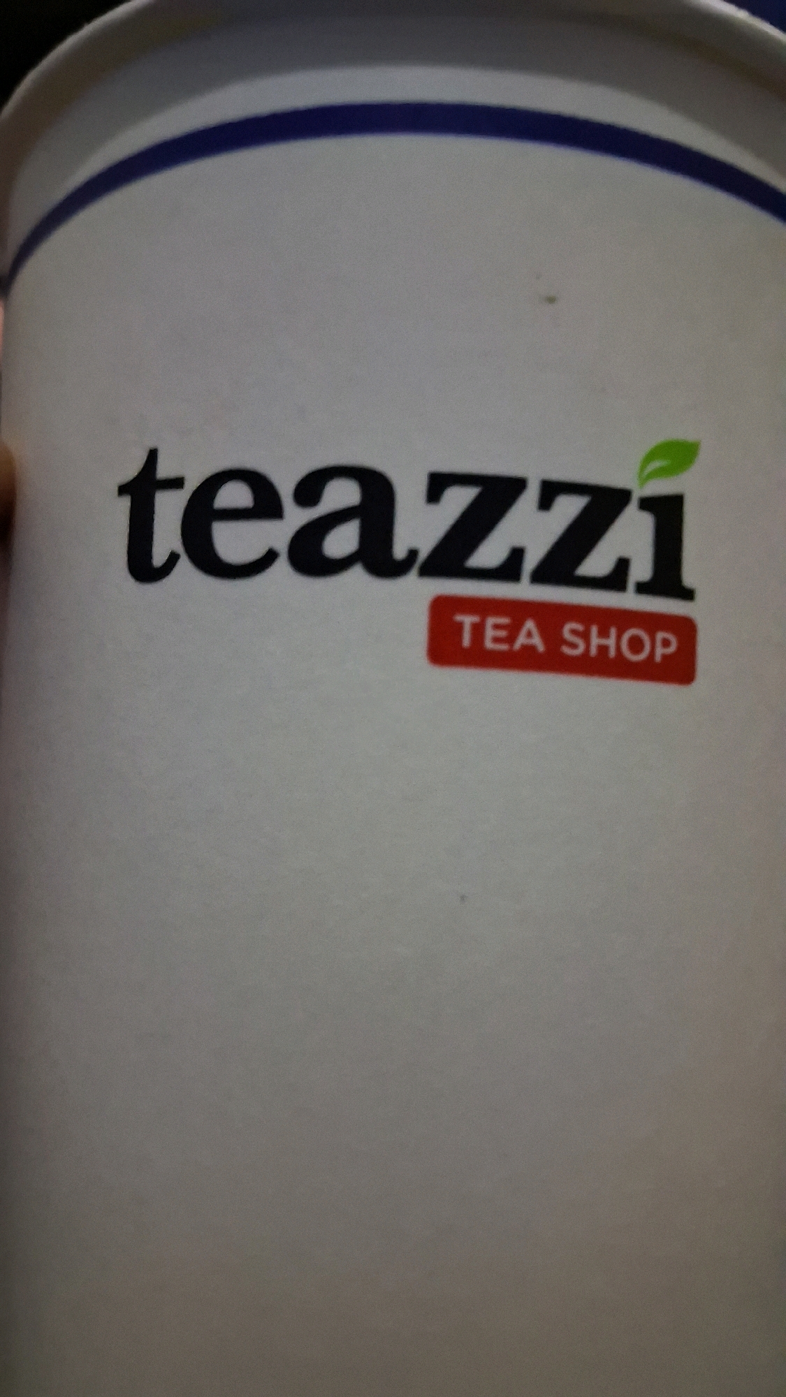 The image shows a close-up of a white paper cup with a blue stripe around the rim. The cup has the logo "teazzi" printed in black, with a small green leaf-shaped design above the letter "i". Below the "teazzi" logo, in a red rectangular box, are the words "TEA SHOP" printed in white. The cup appears to be a disposable to-go cup for beverages. The background is out of focus and appears to be a neutral, indoor setting, possibly a store or cafe. The location in the city of Babakan, Indonesia is not discernible from the image itself.