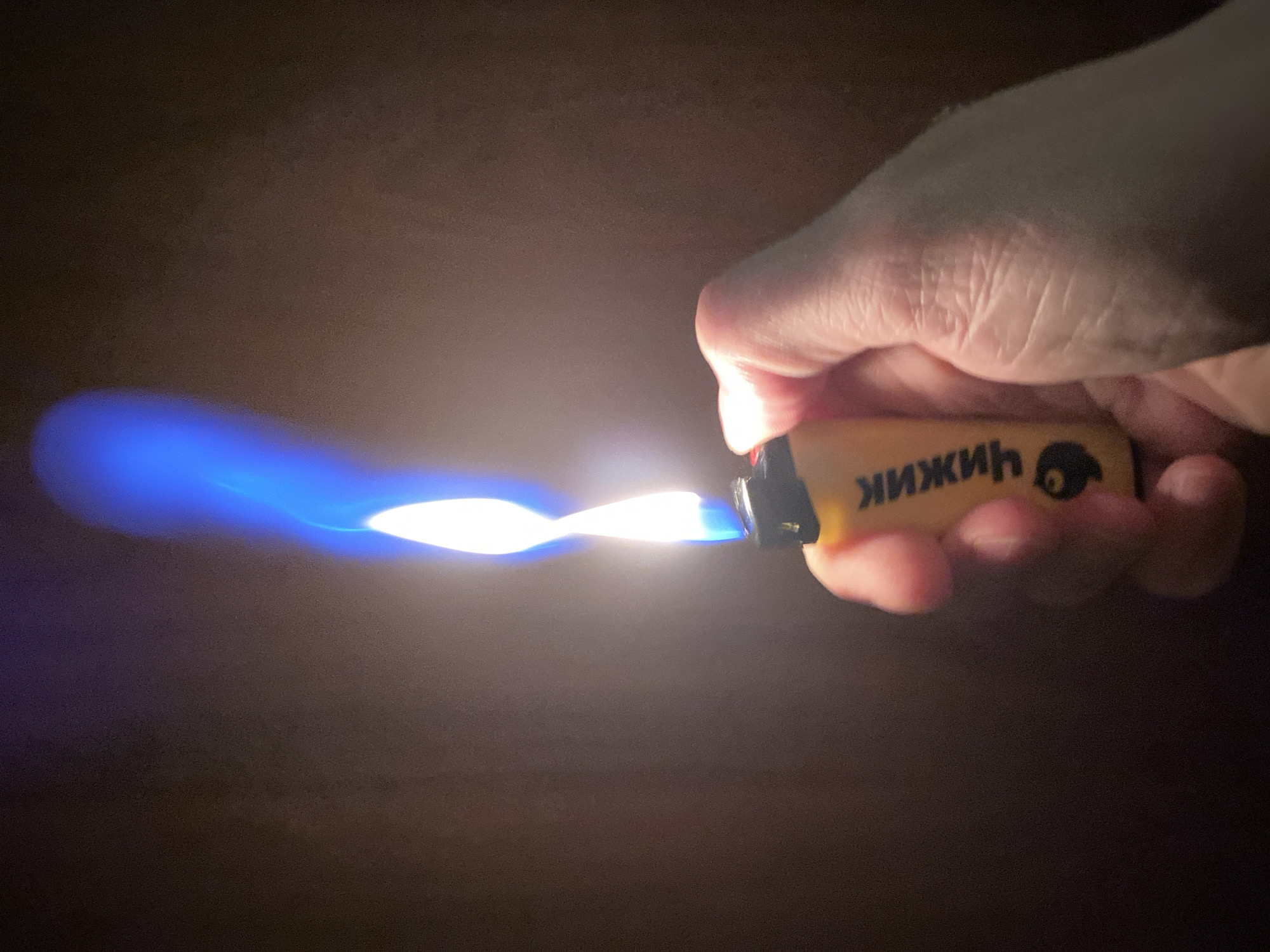A person's hand holds a yellow lighter with a blue flame. The lighter has black accents and text that reads "ЧИЖИК" in black Cyrillic letters, along with a small depiction of a bird. The background is dark and indistinct. The image does not contain any specific location information for Protvino, Russia.