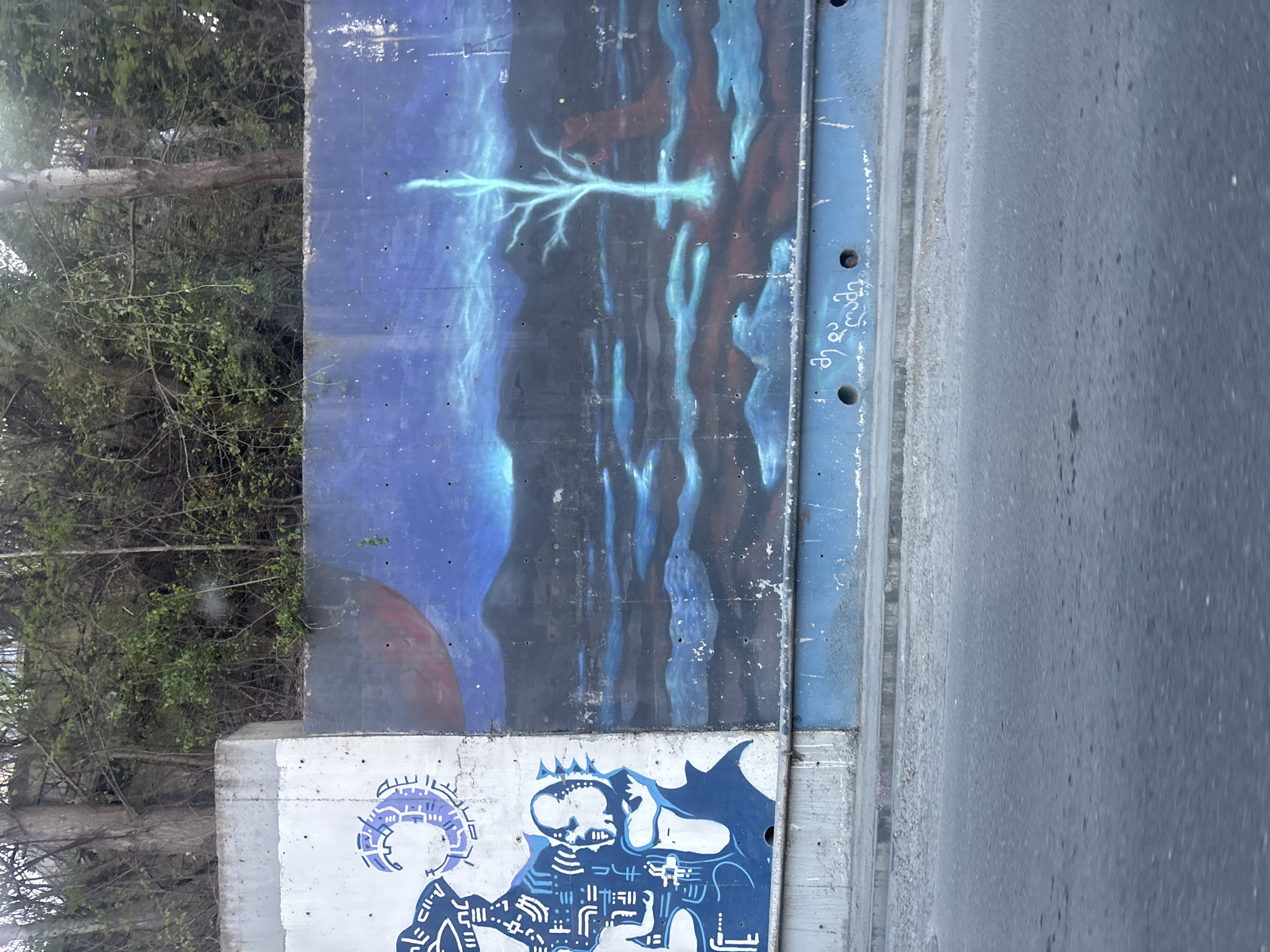 This media file captures a roadside scene in Tbilisi, Georgia, showcasing a concrete wall adorned with two distinct murals. To the left, a dense mass of green trees and branches indicates a natural, possibly overgrown, area next to the structure. On the right, a dark grey asphalt road runs parallel to the wall, suggesting proximity to traffic.
The upper mural, painted on a dark blue background, presents a mystical or cosmic landscape. It features lighter blue and reddish-brown brushstrokes, creating an ethereal ambiance. A prominent, glowing light blue formation, resembling either a tree or a bolt of lightning, stretches horizontally across the center-left. A reddish-brown, semi-circular shape, possibly a planet or sun, is visible towards the lower-left of this artwork.
Below this, on a lighter grey concrete section of the wall, is another mural rendered in blue, black, and white. This artwork depicts stylized figures with a somewhat robotic or skeletal aesthetic. One figure on the left has a complex headpiece, possibly a halo or gear-like design. Another figure features a crowned skull and distinctive sharp, wing-like or cape-like elements, along with intricate patterns that suggest mechanical or futuristic motifs.
A blue metal strip with small drilled holes separates the upper mural from the road. On this strip, white text in Georgian script is visible, spelling "ღმერთო" (Ghmertho), which translates to "God" or "Oh God". The entire scene is brightly lit, indicating it was captured during the daytime.