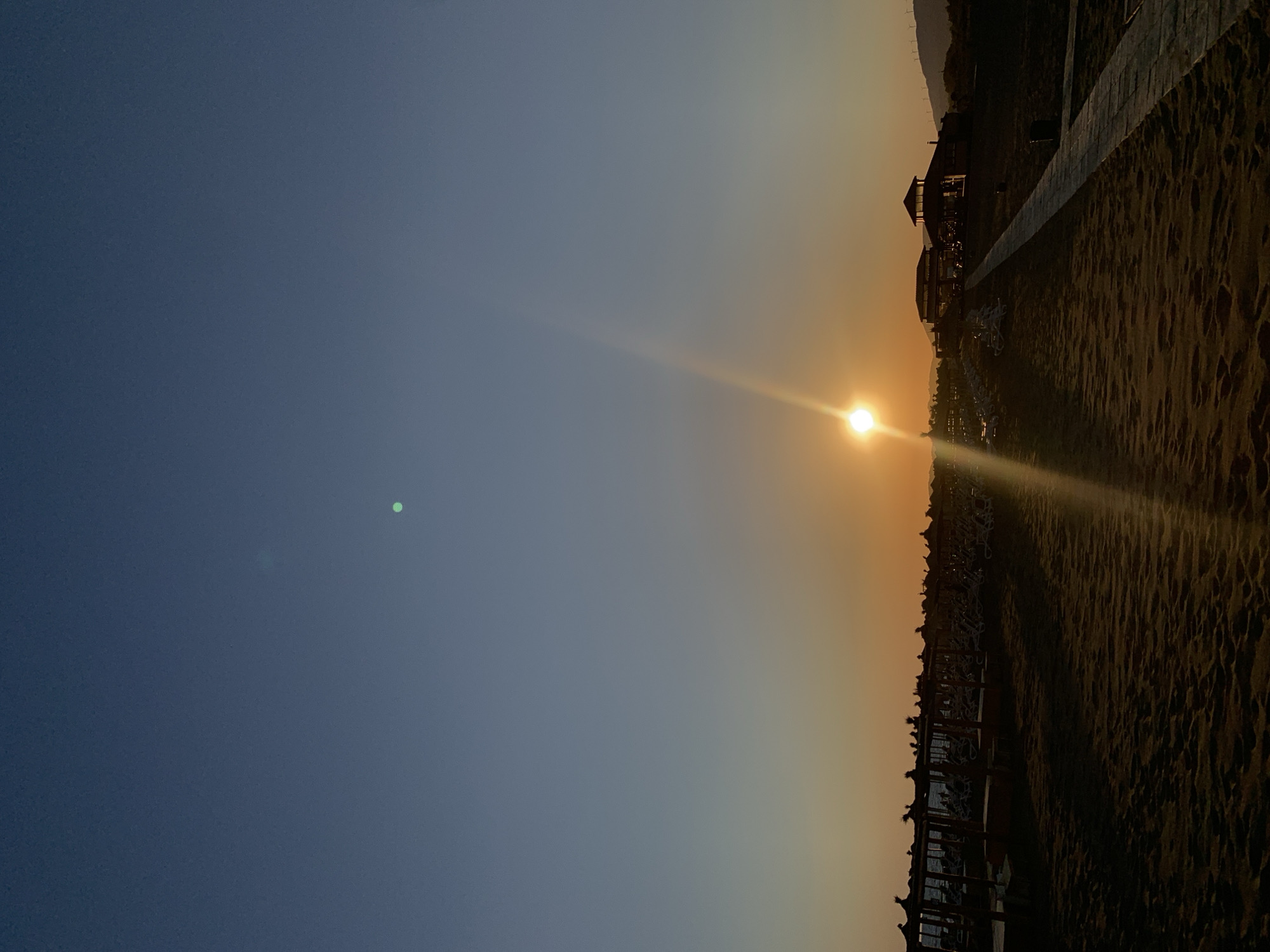 The image depicts a sunset over a beach in Donji Štoj - Shtoji i Poshtëm, Montenegro. The sky transitions from a deep blue at the top to a gradient of orange and yellow as it approaches the horizon. A bright sun, partially obscured by a lens flare, is positioned in the lower half of the frame, casting a strong beam of light upwards and to the left. In the foreground, a sandy beach is visible with subtle rippled textures. A paved walkway extends from the lower right towards a low-rise building with a pitched roof, possibly a restaurant or facility, situated in the middle ground. To the left of the walkway and in the mid-ground, rows of beach chairs and umbrellas are lined up, casting long shadows. The background features a gentle, undulating landscape, with a few wind turbines visible on the hills in the far distance. The overall scene conveys a peaceful and serene end-of-day atmosphere.