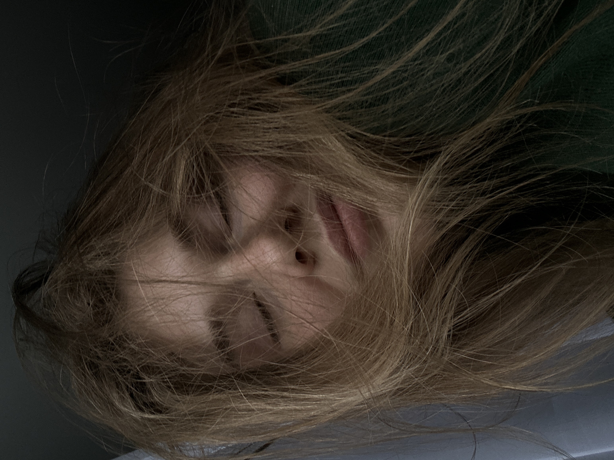 The image shows a close-up of a person's face partially obscured by disheveled, light brown hair. The person's eyes are closed, and the expression is neutral. Only the nose and mouth are clearly visible. The background is dark and indistinct, with hints of a dark green object. The image does not contain sufficient information to identify a specific location within Kazan, Russia or to determine any actions or interactions taking place.