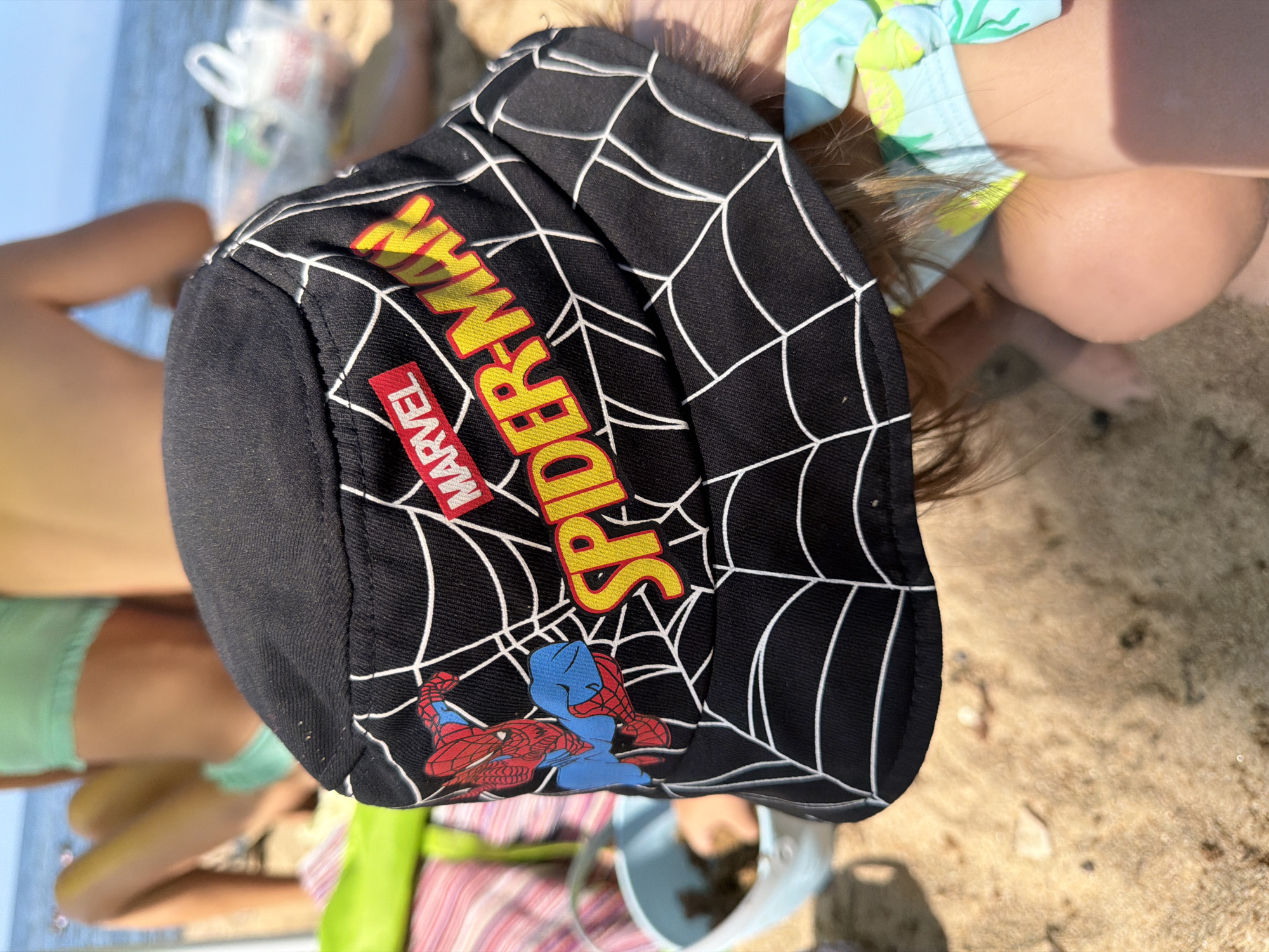 A child wearing a black bucket hat with a Spider-Man design and web pattern sits on a sandy beach near Maksimiha, Russia. The hat is adorned with the "MARVEL" logo and the words "SPIDER-MAN" in yellow lettering. The child's face is obscured by the hat. In the background, other children are visible, along with beach accessories and the sea. The environment is a beach, characterized by sand and water, with the sky in the distance.