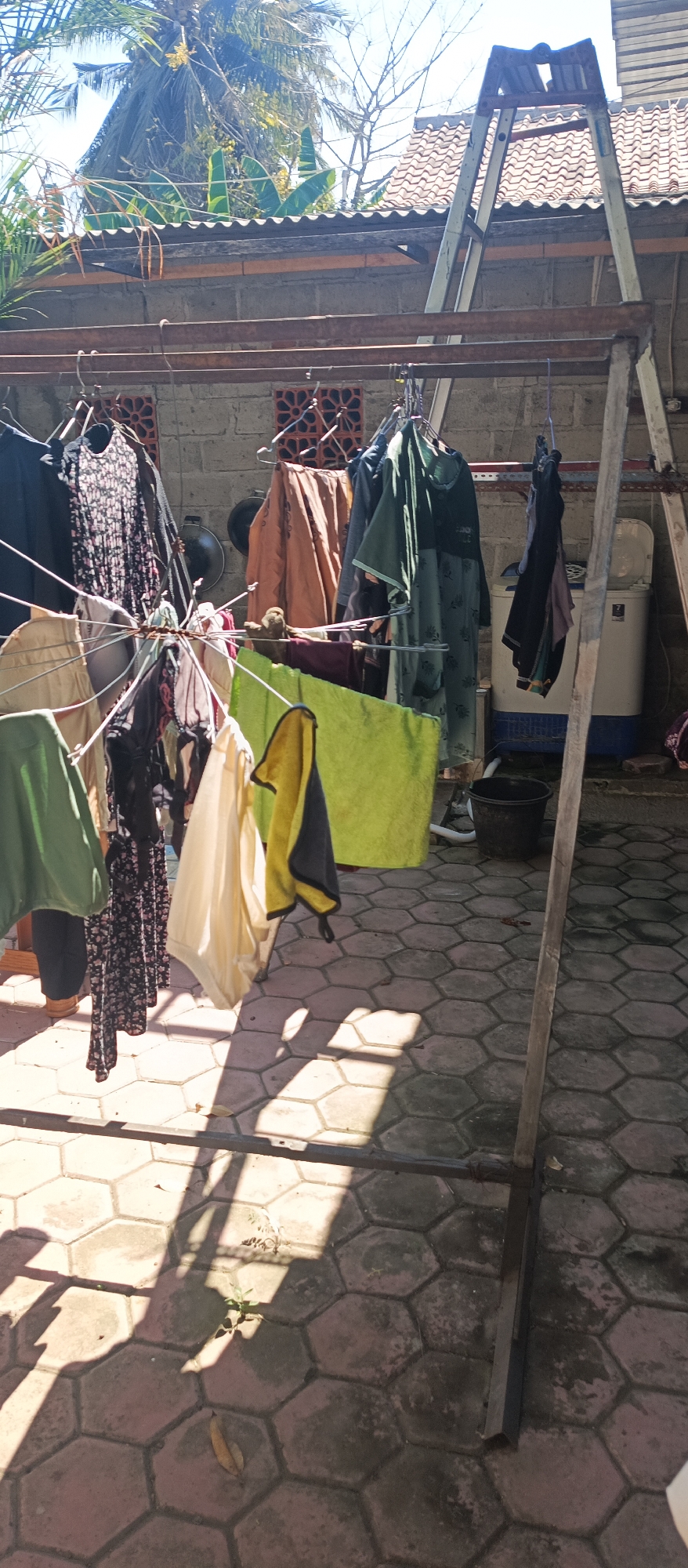 An outdoor scene in Jatisari, Indonesia, features a clothesline laden with various garments, including dresses, shirts, and towels. The clothes are hung on metal racks under a corrugated metal roof structure, with a brick wall visible in the background. Sunlight casts strong shadows across the hexagonal patterned tiled ground. A washing machine and a black bucket are also present in the scene. Tropical foliage, including palm trees and banana leaves, is visible overhead.