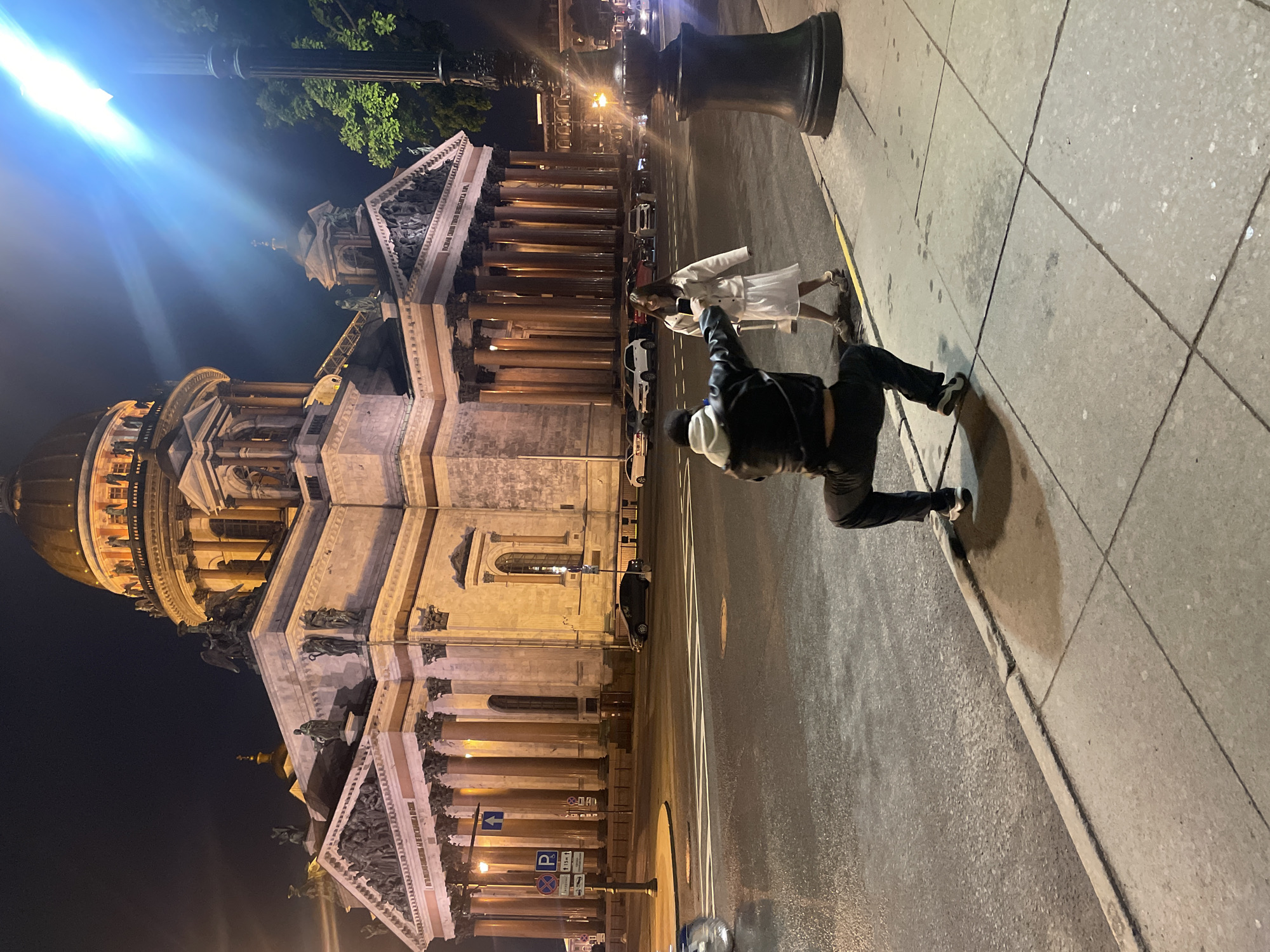 At night in Saint Petersburg, Russia, two people stand next to a street and sidewalk beside Saint Isaac's Cathedral. One person, wearing black pants and a black jacket, is on the sidewalk pointing towards the cathedral. The other person, wearing a white dress or jacket, stands next to them facing the cathedral. The cathedral is lit up and is in the background. There are street lights and traffic on the street between the cathedral and the sidewalk. A street sign and a parking sign are visible.