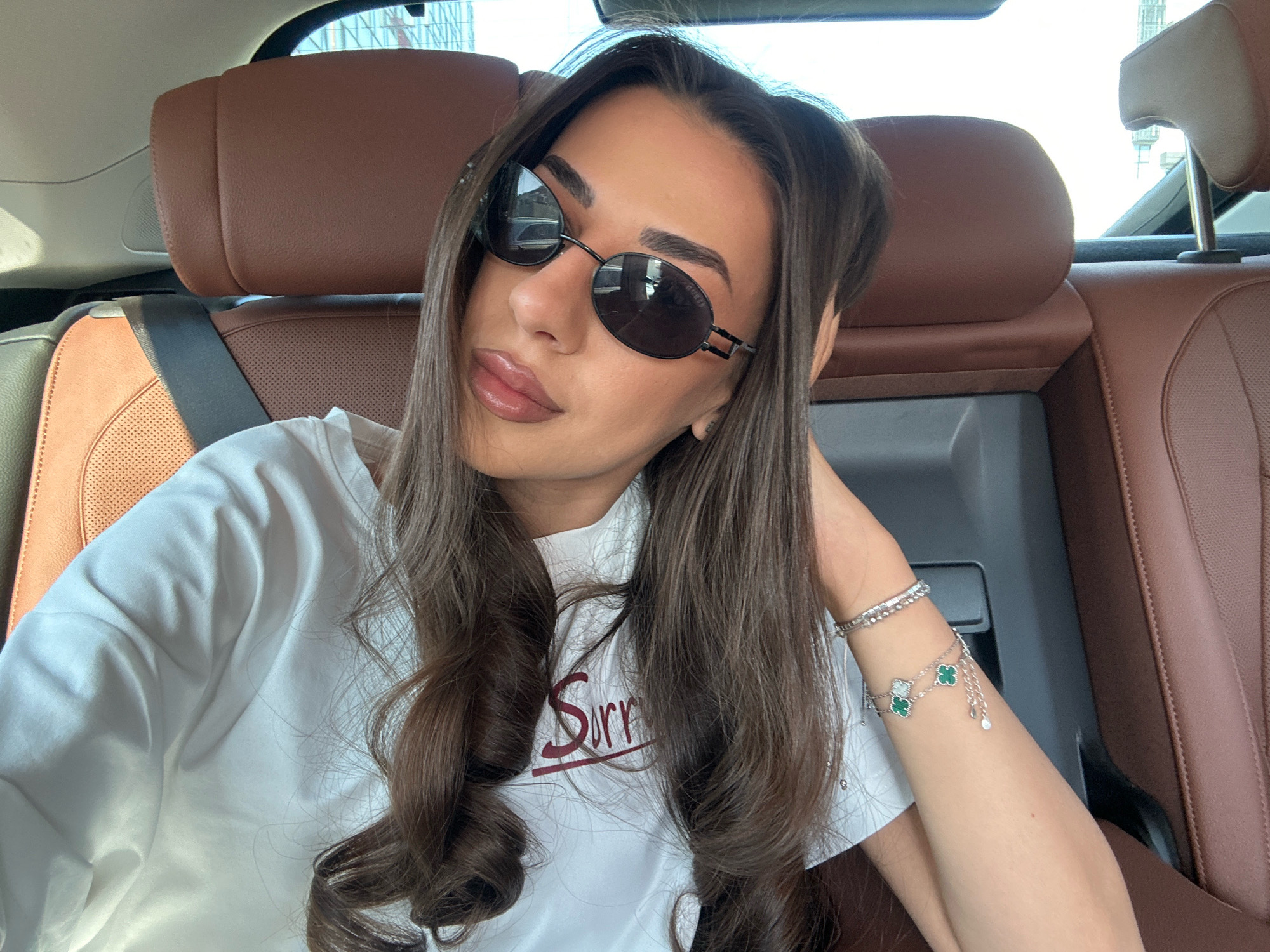 A young woman with long, dark brown hair is seated in the back of a car. She is wearing large, oval sunglasses and a white t-shirt with a red graphic. Her left arm is resting on her head, and she is wearing a bracelet with green clover-shaped charms. The car's interior is visible, with brown leather seats. The background shows a cityscape through the car windows, suggesting an urban environment. The image appears to have been taken in the city of Sofia, Bulgaria.