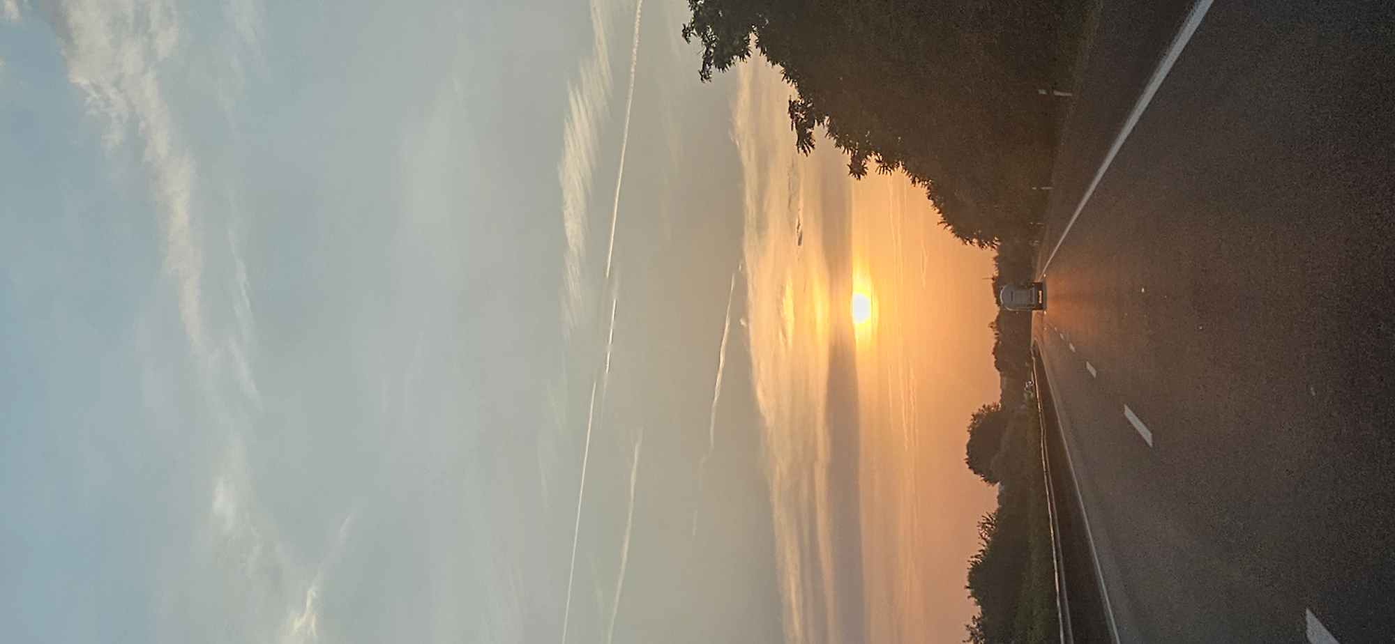 The image captures a sunset over a highway in Balatonszabadi, Hungary. The sun, a bright orange orb, is low in the sky, partially obscured by wispy clouds. The sky transitions from a pale blue at the top to shades of orange and yellow near the horizon, reflecting the warm glow of the setting sun. Several white contrails are visible in the upper portion of the sky.
The highway stretches forward, with a single lane visible in the foreground, marked by white lines. The road surface is asphalt, appearing dark and wet in places, reflecting the sunset's light. On the right side of the road, a dense line of trees and bushes creates a dark silhouette against the vibrant sky. On the left side, a guardrail separates the highway from a grassy embankment, beyond which more trees are visible.
A white truck with a cylindrical tank is traveling away from the viewer on the highway. It is illuminated by the setting sun, casting a long shadow on the road. Other vehicles are faintly visible in the distance on the highway. The overall scene depicts a serene, yet dynamic, moment on a rural highway at dusk.
