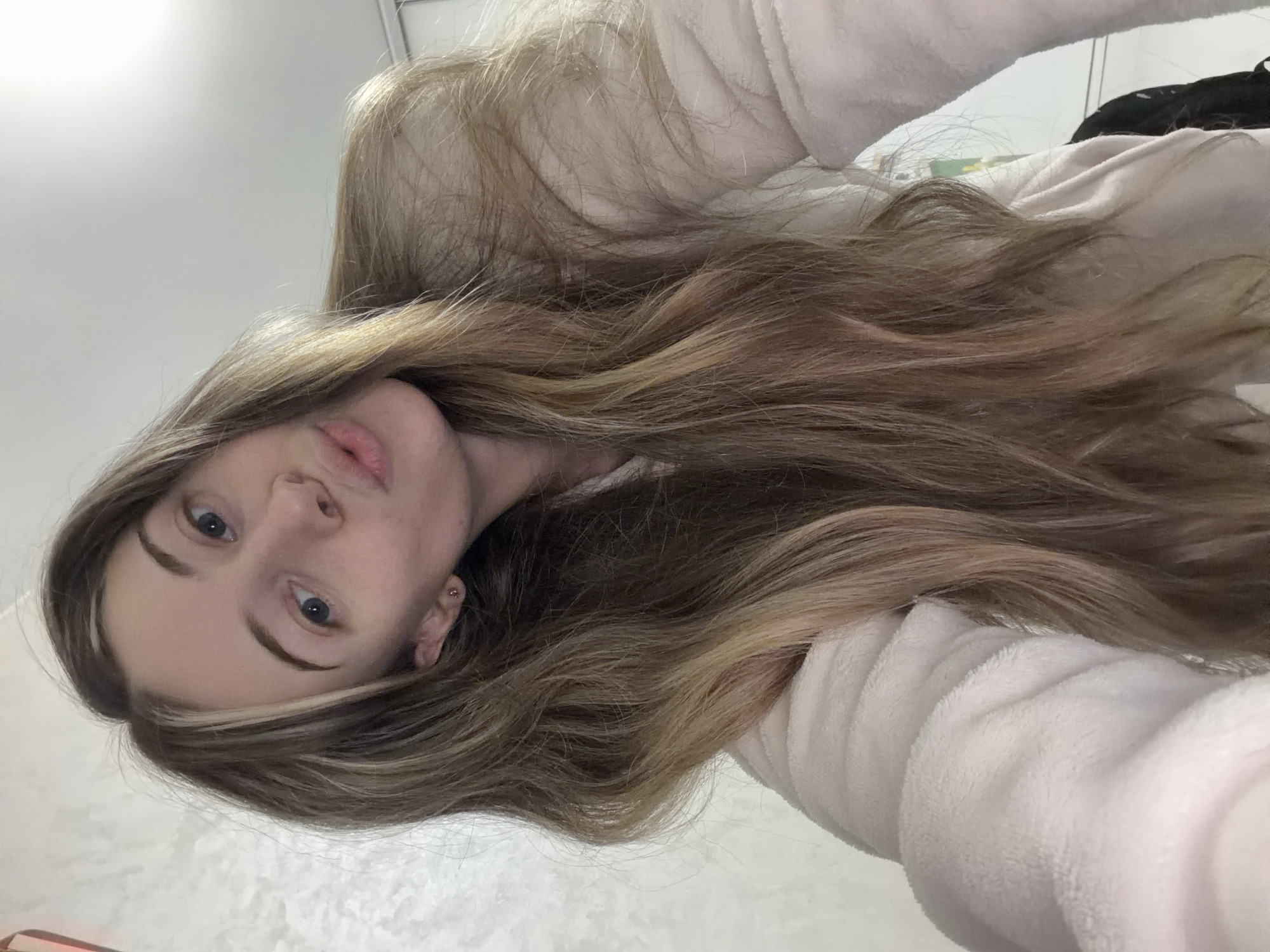 A young woman with long, light brown hair, styled with darker roots and lighter highlights, is facing the camera. She has fair skin, blue eyes, and distinct eyebrows. She is wearing a soft, light-colored garment with a textured fabric, which could be a robe or sweater. The background is a neutral-toned wall, possibly white or light gray. The photo appears to be taken indoors, likely using a selfie camera. There is no information given that indicates the location is in the city of Plovdiv, Bulgaria.