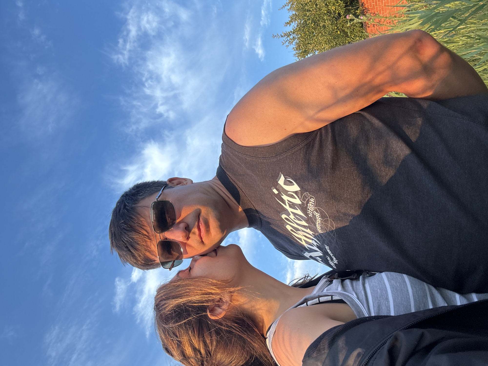 A woman is kissing a man on the cheek in an outdoor setting. The man is wearing sunglasses and a dark t-shirt with a graphic print, and he is looking directly at the camera. The woman is leaning into the man, with her hair partially obscuring her face. The background features a blue sky with scattered white clouds, and a hint of greenery and a brick wall on the right side. The lighting suggests it is either early morning or late afternoon. The specific location in the city of Karachunivka, Ukraine, is not discernible from the image itself.