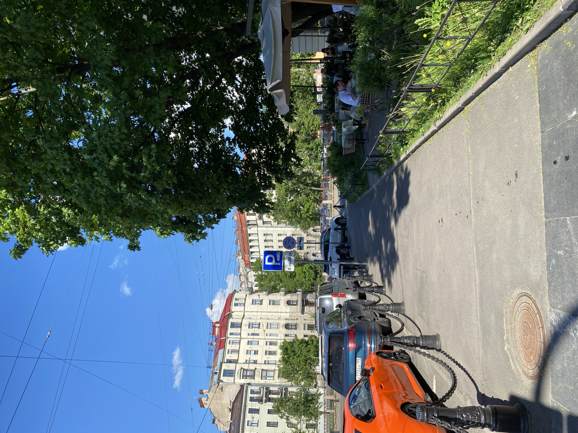 The image depicts a sunlit street scene in Saint Petersburg, Russia, featuring a sidewalk, an outdoor cafe, parked cars, and architectural buildings under a clear blue sky.
On the left, large, leafy green trees provide shade over a section of the sidewalk where an outdoor cafe is set up. Several people are seated at tables and chairs, some under a large white awning, appearing relaxed and enjoying the sunny weather. A metal fence with green foliage and yellow dandelions separates the cafe area from the street.
Along the right side of the street, multiple cars are parked. Prominently visible are a bright orange sports car and a blue SUV bearing the Russian license plate "577OA198" (indicating the Saint Petersburg region). The cars are secured by decorative black bollards linked with chains. A blue parking sign with a white "P" symbol clearly indicates paid parking hours from "8.00-20.00". Overhead utility wires are visible against the sky.
In the background, multi-story, light-colored buildings with red-tiled roofs, characteristic of Saint Petersburg's architecture, line the street. A circular metal manhole cover is visible on the pavement, featuring Cyrillic text, including "ГОРВОДОКАНАЛ" (Gorvodokanal - city water utility), "С-Пб" (S-Pb, an abbreviation for Saint Petersburg), and the year "2010".
The scene suggests a pleasant day, likely mid-morning or afternoon, given the bright sunlight and visible shadows. People are engaged in leisurely activity at the cafe in a bustling urban setting.
