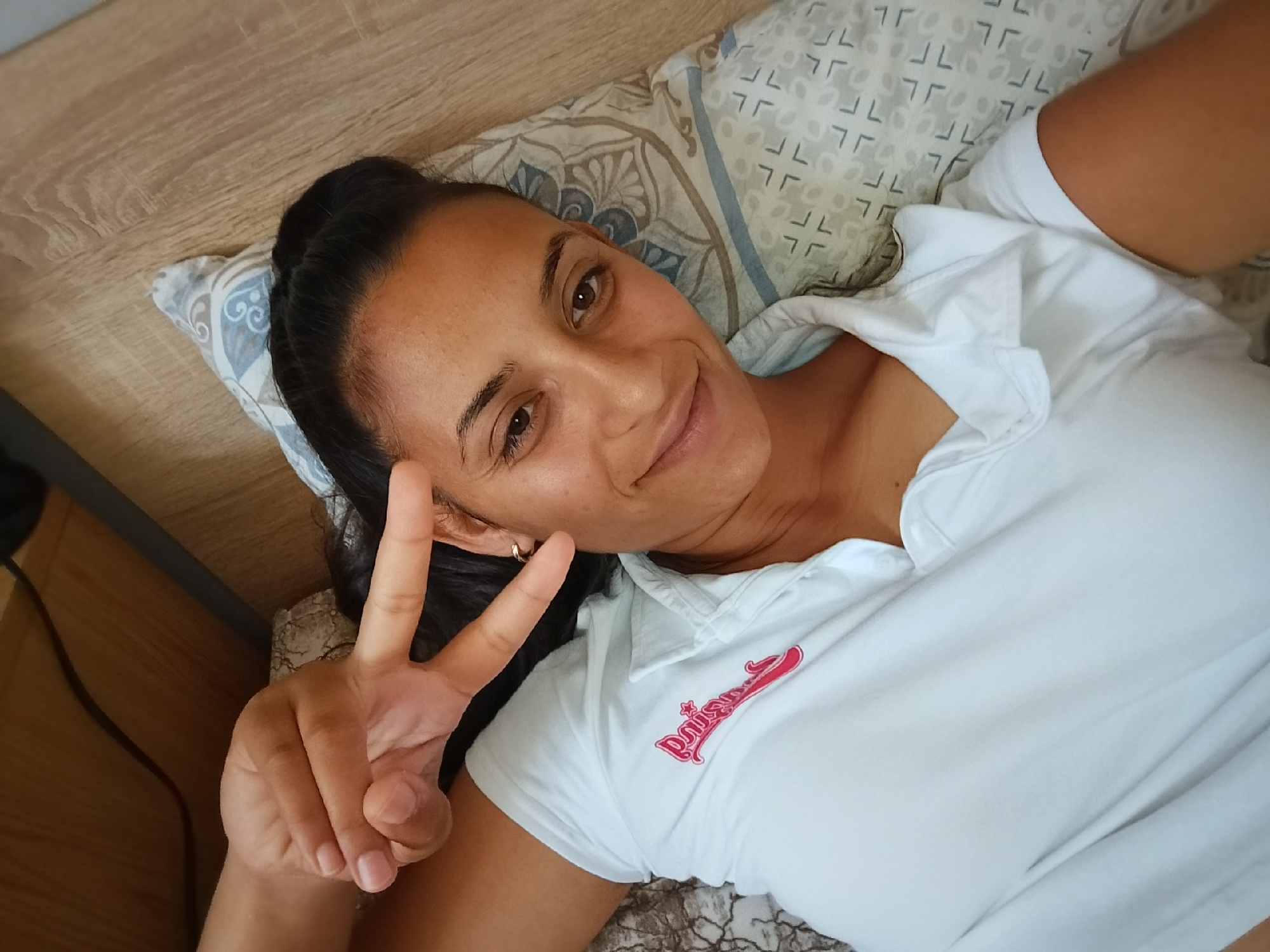 A high-angle, medium shot captures a woman lying on a bed, holding up two fingers in a peace sign. She has dark hair pulled back, brown eyes, and is wearing a white collared t-shirt with a pink logo. The bed has a patterned pillow and a wooden headboard. The woman appears to be in a domestic setting.