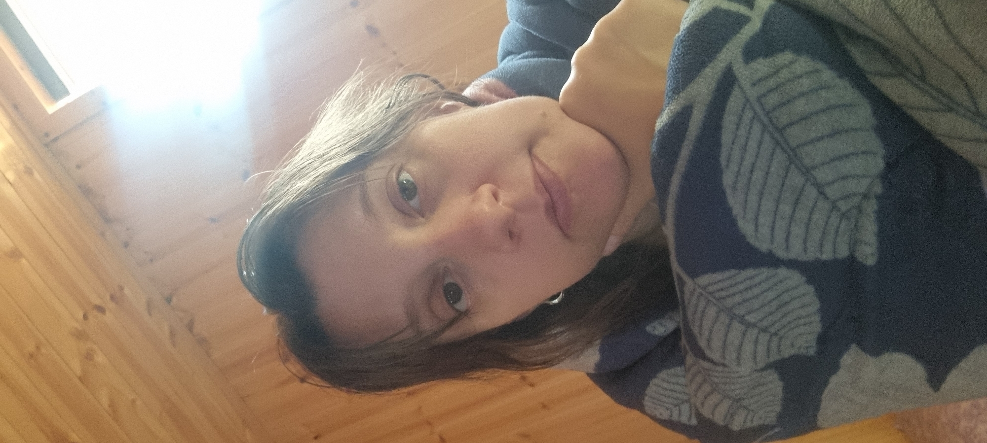An image captured from a low angle features a young woman indoors, likely reclining or lying down, looking towards the camera with a calm, slightly smiling expression. Her dark hair frames her face, and her eyes are open. She is partially covered by a blue fabric adorned with a repeating pattern of light-colored leaves.
The setting appears to be a cozy room with extensive wooden paneling on the walls and ceiling, suggesting a rustic or cabin-like interior. A bright window or skylight is visible in the upper left portion of the image, emitting intense natural light that illuminates the scene and creates a warm glow. The strong light indicates it is daytime.
The woman's posture and the soft fabric she is wrapped in convey a sense of comfort and relaxation. No visible text is present in the image. Given the context of Dunnae-myeon, South Korea, the scene depicts a moment of quiet repose in a domestic setting within that region.