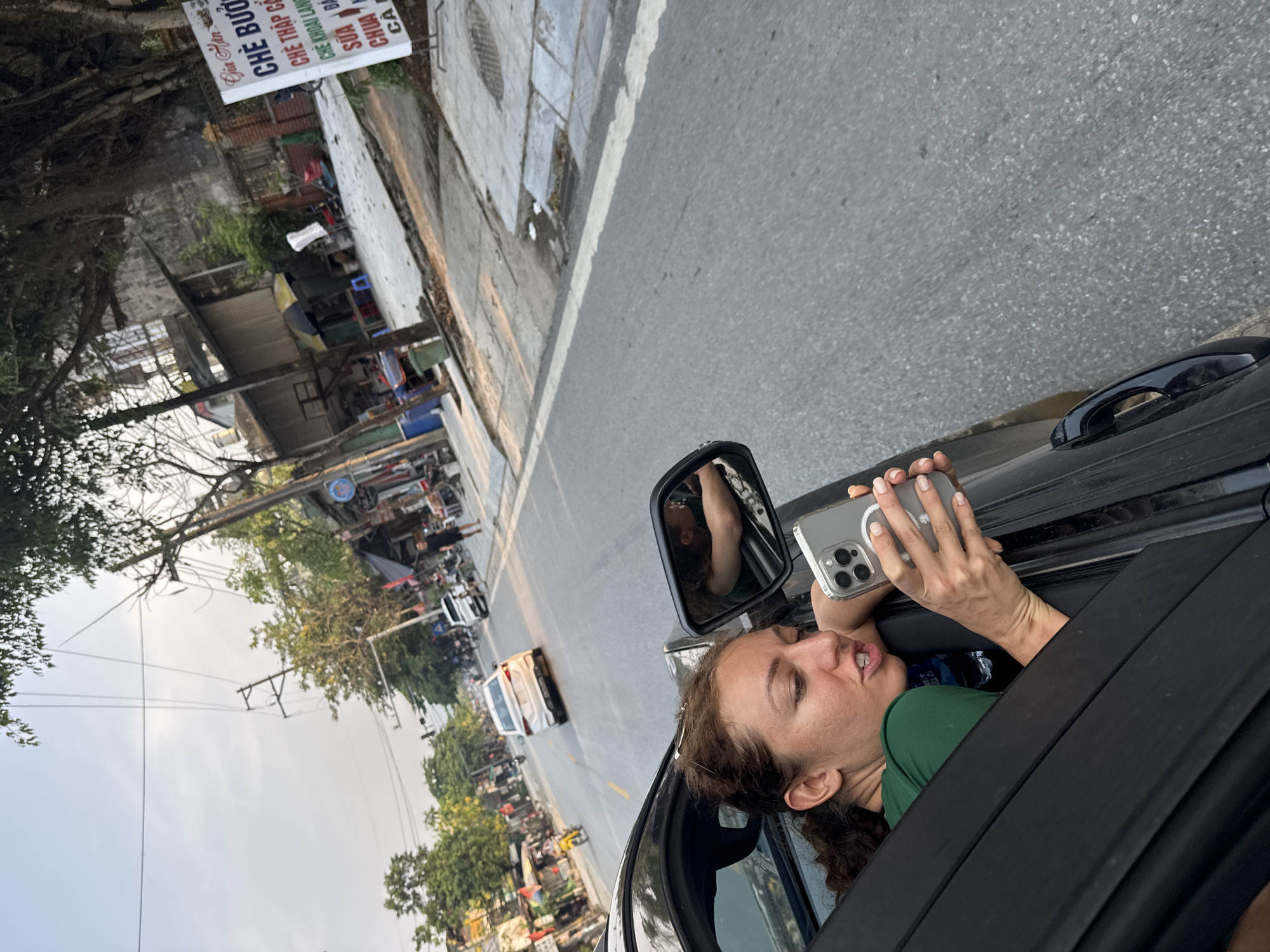 The image, taken in Tân Quang Commune, Vietnam, captures a candid moment of a woman leaning out of a car window on a bustling street. The perspective is angled, with the car door and window framing the foreground and the street extending into the midground.
The main subject is a woman with medium-brown, wavy hair, dressed in a dark green top. She leans out of the car, resting her right arm on the window frame, her gaze directed forward and slightly downward, with a neutral, slightly open-mouthed expression. She holds a silver or light grey smartphone, likely an iPhone with its distinctive triple-lens camera, in both hands, positioned as if taking a photo, recording, or viewing its screen. Her reflection is visible in the car's side mirror. A clear plastic water bottle is also seen inside the car.
The scene depicts a vibrant street during daytime, under soft, diffused lighting. An asphalt road runs alongside a concrete sidewalk, complete with a visible manhole cover. Along the roadside, numerous small buildings, possibly shops or market stalls, are present, some featuring awnings. The street is active with various vehicles, including several motorbikes, a white van, and another white car in the distance. Utility poles with a web of wires stretch across the sky, while abundant green trees and foliage line the street.
A notable detail is a white sign with blue Vietnamese text on the left side of the road. The top line reads "Gia Hân." Below it, several Vietnamese sweet desserts (Chè) are listed, including "CHE BUOC," "CHÈ THẬP CẨM," "CHE KHOAI LANG" (sweet potato che), and "SƯA ĐẬU" (soy milk). The image conveys a sense of observation and engagement with the local street environment from within a moving or stationary vehicle.