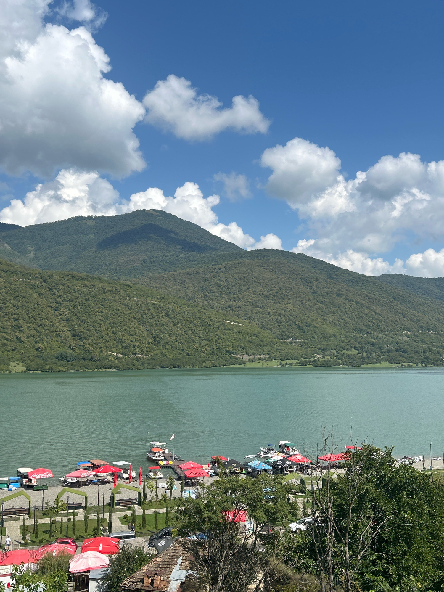 The image depicts a scenic view of the Zhinvali Reservoir near the city of Shalikiantkari, Georgia. The reservoir's turquoise water is bordered by lush, green mountains. The foreground shows a lively lakeside scene with several small boats moored and docked. Along the shore, there are numerous red umbrellas, some of which appear to be part of outdoor seating areas for restaurants or cafes. Several cars are parked nearby, and a few people can be seen milling around the area. The sky is a vibrant blue, dotted with fluffy white clouds, contributing to the overall picturesque atmosphere. The scene suggests a popular recreational spot, likely during the daytime on a clear day.