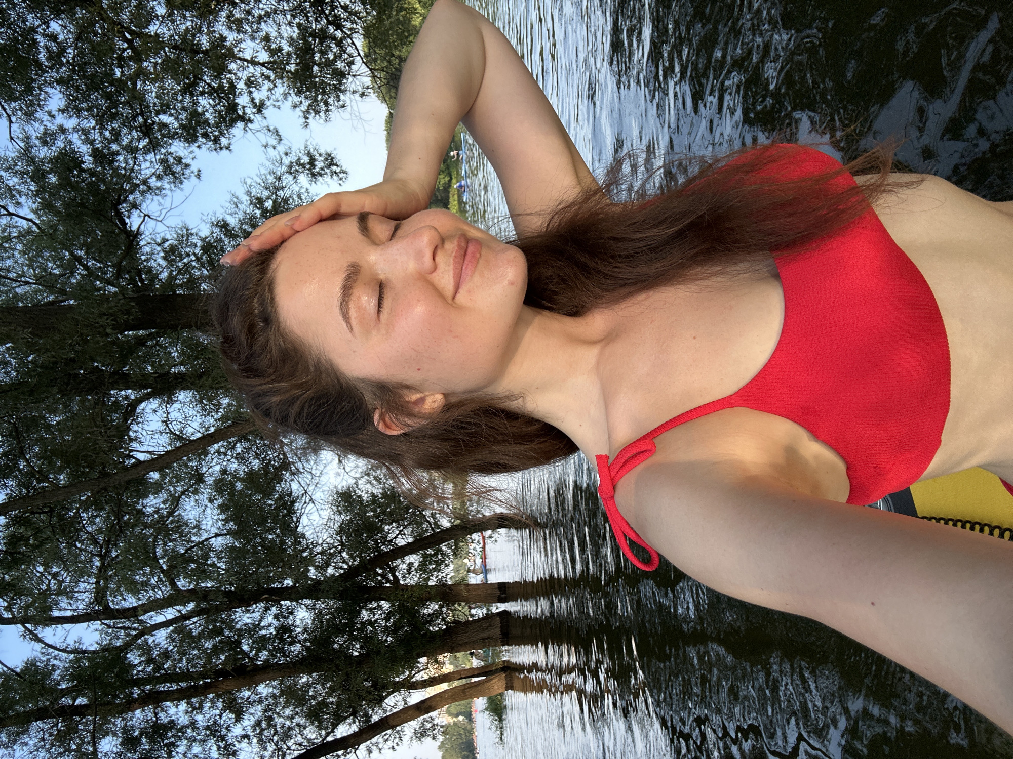 A young woman with long brown hair and closed eyes is taking a selfie in the Ufa River in Ufa, Russia. She is wearing a red bikini top and has her right hand on her forehead. The water is dark and rippling, with trees on the left and in the background. The sky is blue and clear. There are other people on the water in the distance, some on paddleboards.