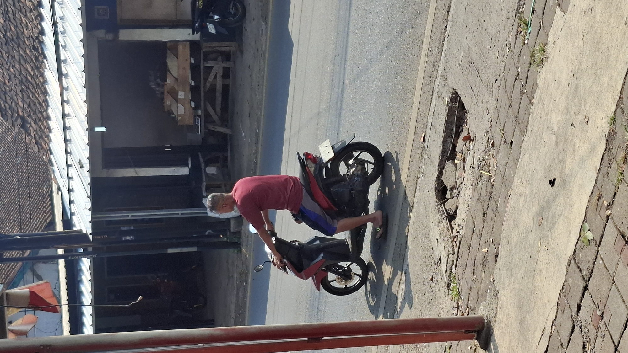 A man with short, graying hair is riding a red and black scooter on the right side of a paved road. He is wearing a maroon t-shirt and blue and gray striped shorts. His feet are bare, wearing flip-flops. The scooter has a license plate that is not clearly legible. He is casting a shadow to his left. In the background, a storefront with a corrugated metal roof is visible, with displays of goods on wooden tables. To the left of the storefront, a red pole stands on the sidewalk. The sidewalk is made of interlocking pavers and concrete sections. A large pothole with exposed rebar and debris is present in the sidewalk near the road. The overall environment appears to be an urban or semi-urban setting in Cilaku, Indonesia, likely a commercial area.