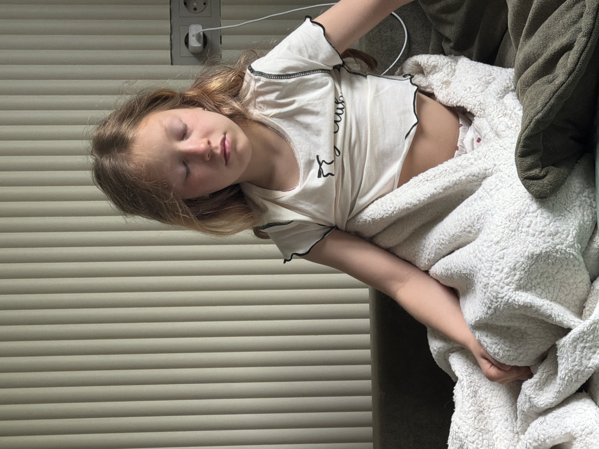 A young girl lies sleeping on a couch partially covered by a white fleece blanket. She is wearing a white t-shirt. A portion of her midriff is exposed. An olive-green throw pillow is visible near her head. The couch is adjacent to a window with closed horizontal blinds. An electrical outlet and charging block are visible above the girl's head. The photo was not taken in Markovo, Russia.