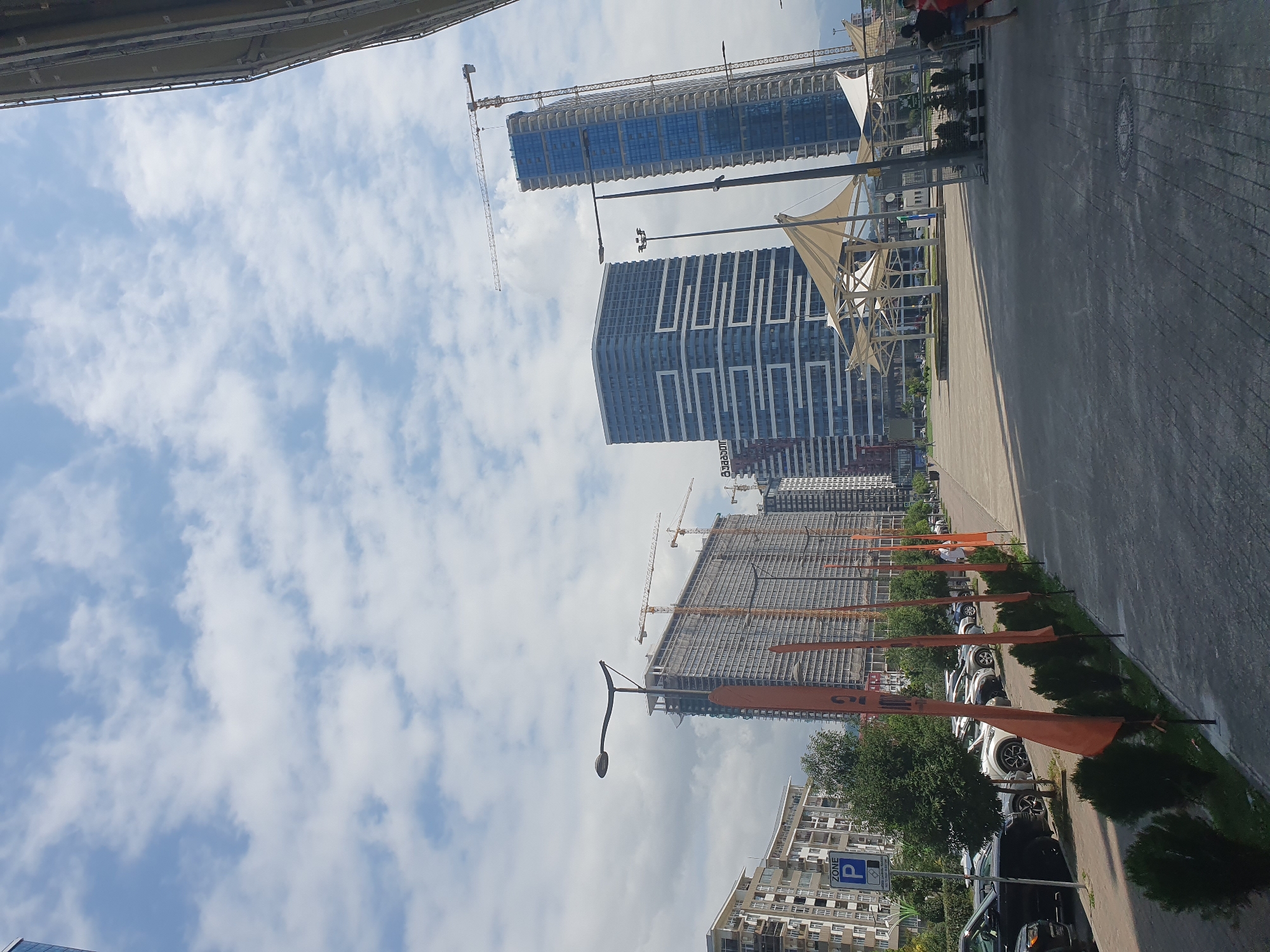 This is a daytime, eye-level, full shot photograph taken in Batumi, Georgia, likely near the Batumi Boulevard. The image captures a street scene with several modern high-rise buildings under construction and recently completed. In the foreground, a paved walkway runs along the right side, with a line of small green bushes and orange flags planted in the ground. To the left of the walkway is a parking lot filled with cars, and beyond that, several apartment buildings.
The background is dominated by tall buildings. One particularly striking building is a blue skyscraper with a reflective facade. Adjacent to it is a taller building that appears to be under construction, with cranes visible at the top. Further to the left, another large residential building is under construction, also with cranes. In the middle distance, there is an open area with white, sail-like structures, possibly a canopy or part of a public space.
The sky is partly cloudy, with scattered white clouds against a blue backdrop. The lighting suggests it is a bright, possibly sunny day. A few people are visible in the scene: two individuals, one in a red top and the other in a dark top and shorts, are walking away from the camera on the right side of the walkway. Another person is visible standing further down the walkway. There is a parking sign on the left side of the image. The overall impression is one of urban development and modern architecture in a coastal city.